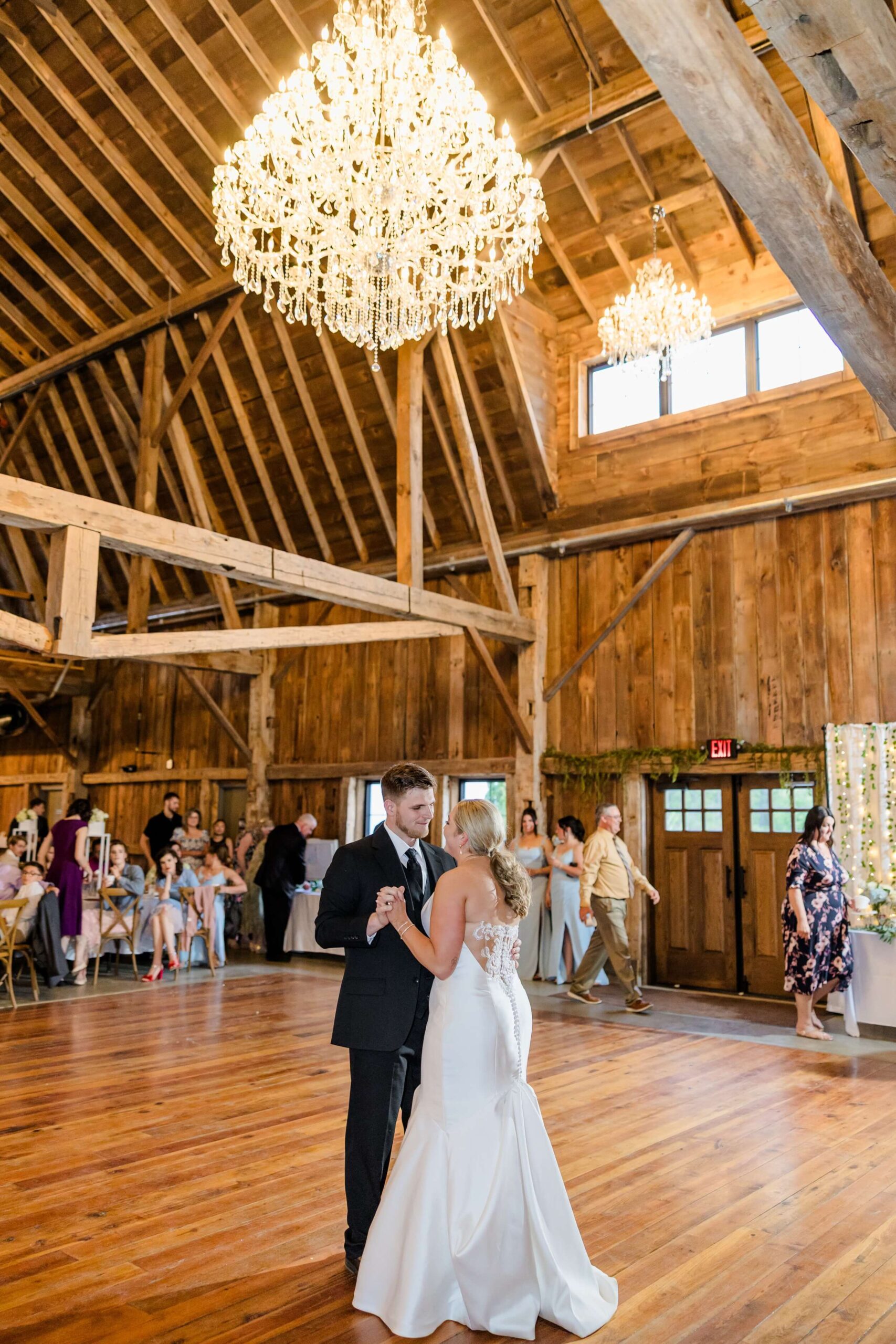 Newlyweds dance under large chandeliers in an exposed beam barn on the dance floor at The Glenn Royal Barn Venue