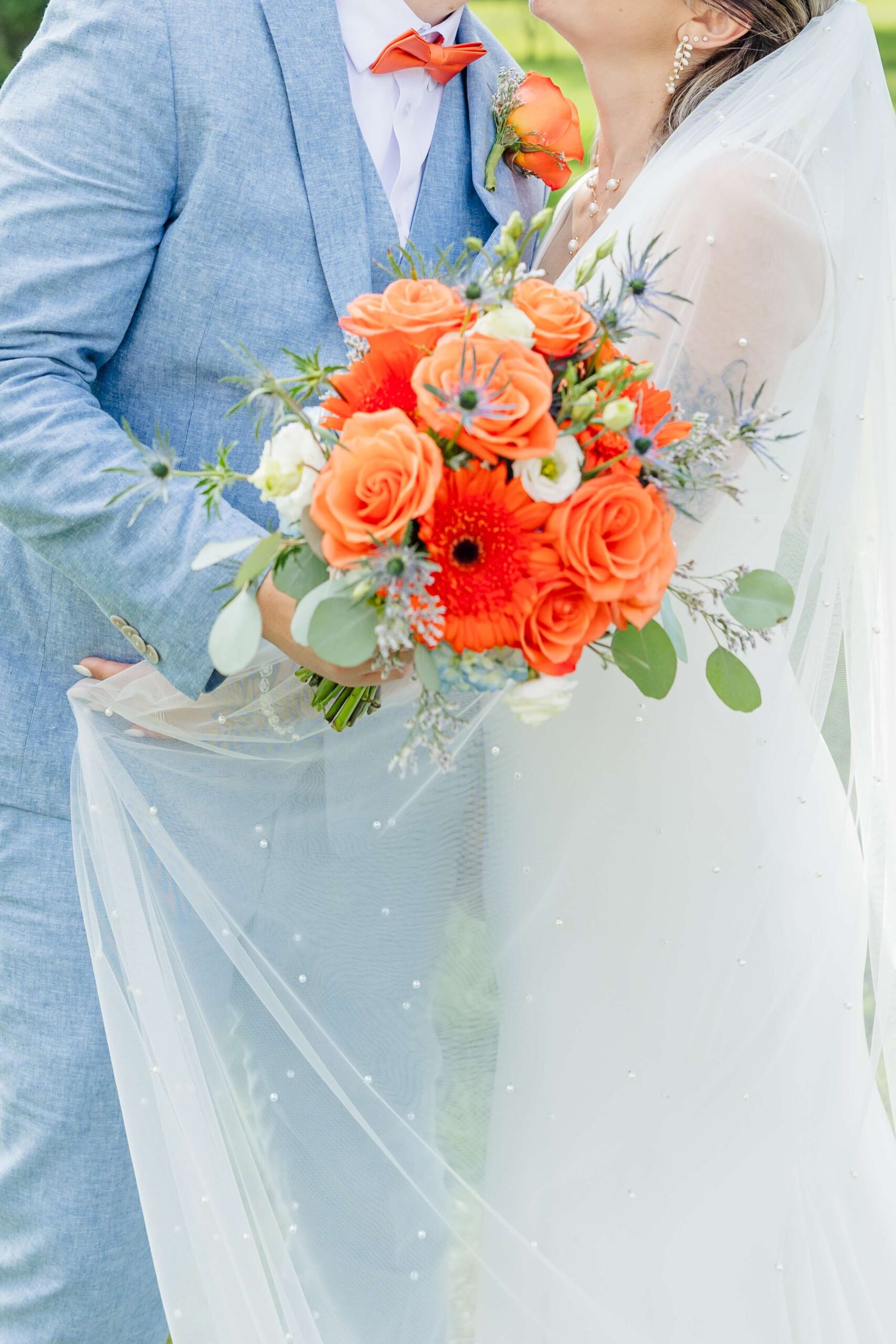 Details of a bride's orange bouquet being held by the groom in a light blue suit while they hug