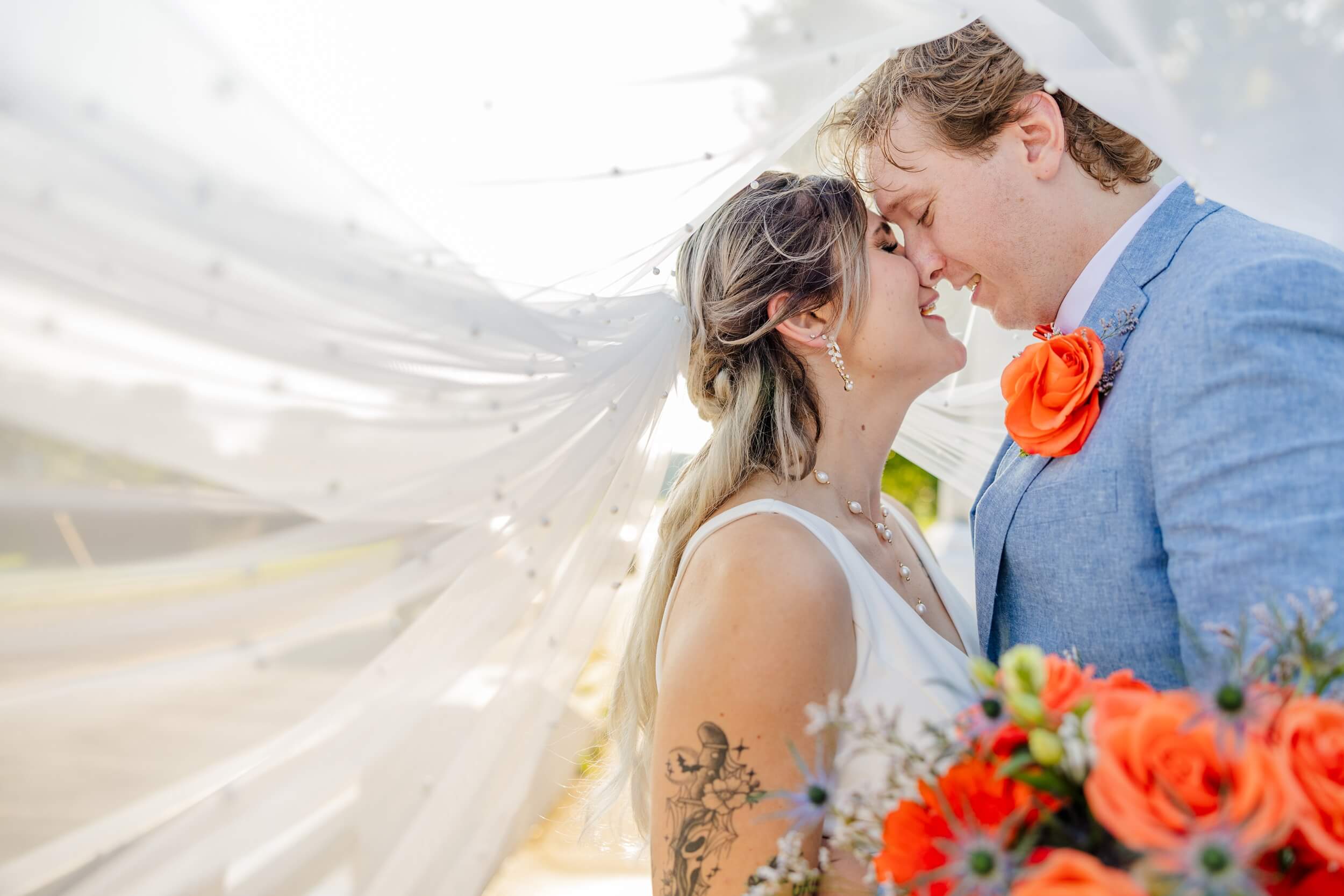 A happy bride with pearl jewelry and her groom in a blue suit lean in for a happy kiss while hiding under the veil with orange florals at their Planterra Conservatory wedding
