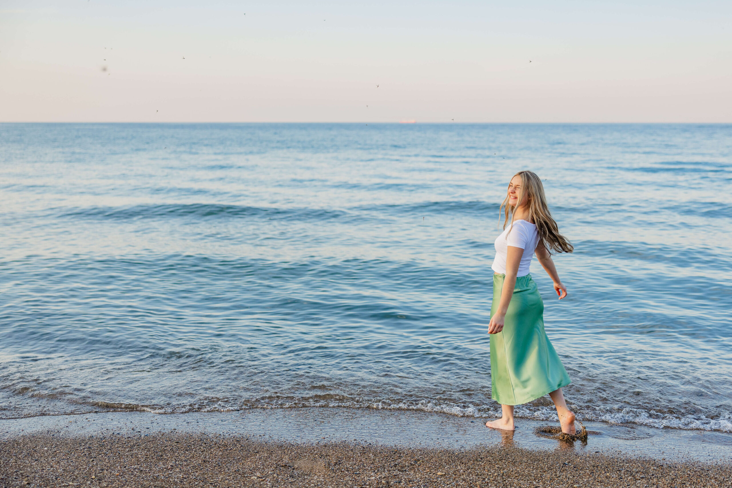 A high school senior in a white shirt and green skirt dances by the water at sunset
