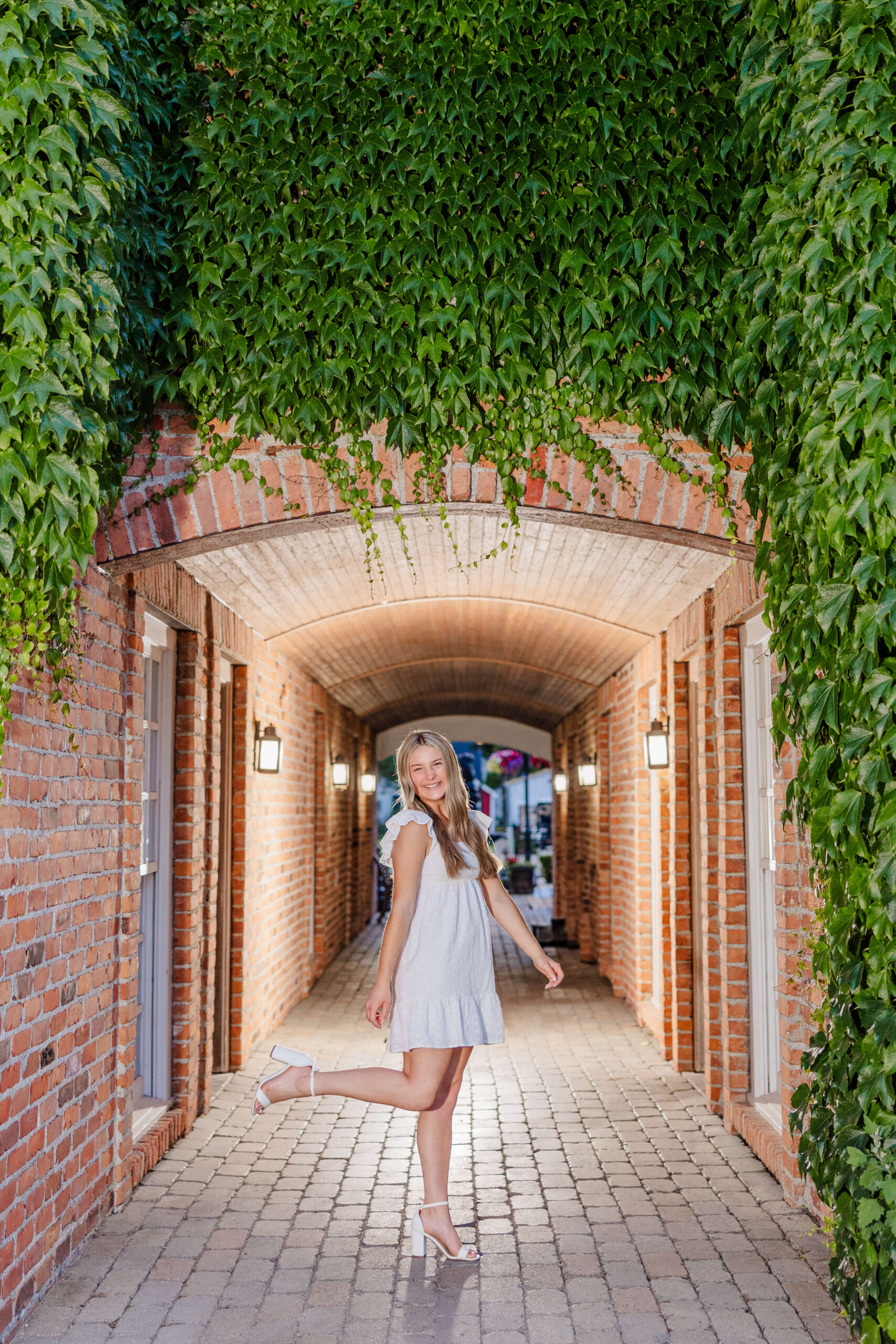 A happy high school senior pops one leg while standing in a brick hallway covered in ivy in a white dress