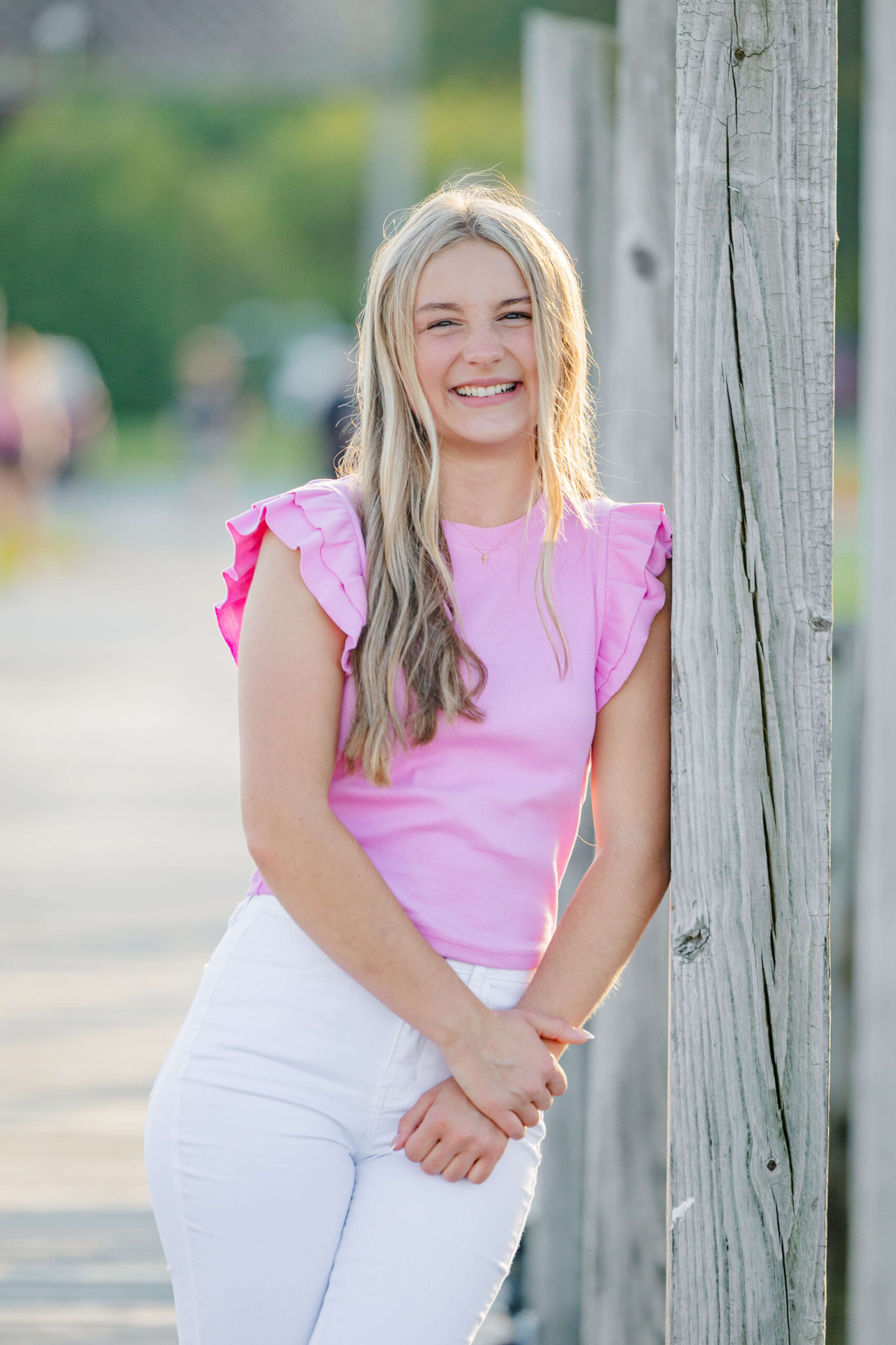 A high school senior in a pink blouse and white pants leans on a wooden post in a park at sunset after visiting boutiques in michigan