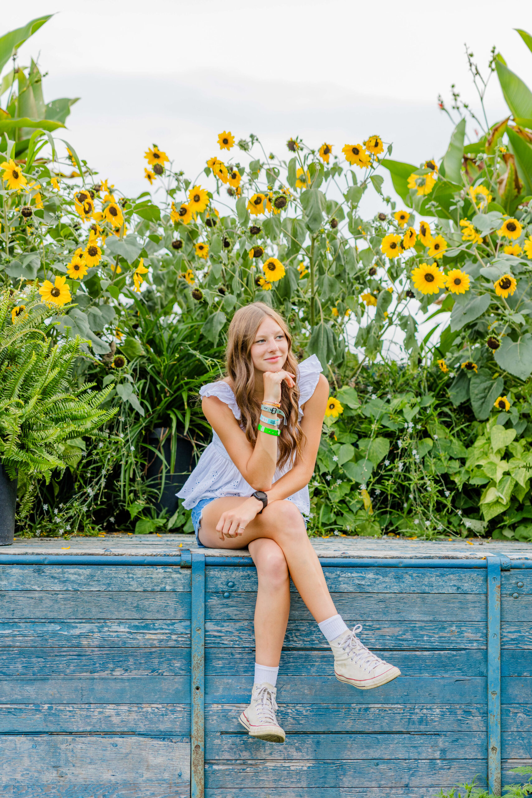 A teenage girl sits on a wooden wall thinking surrounded by yellow flowers in a white shirt