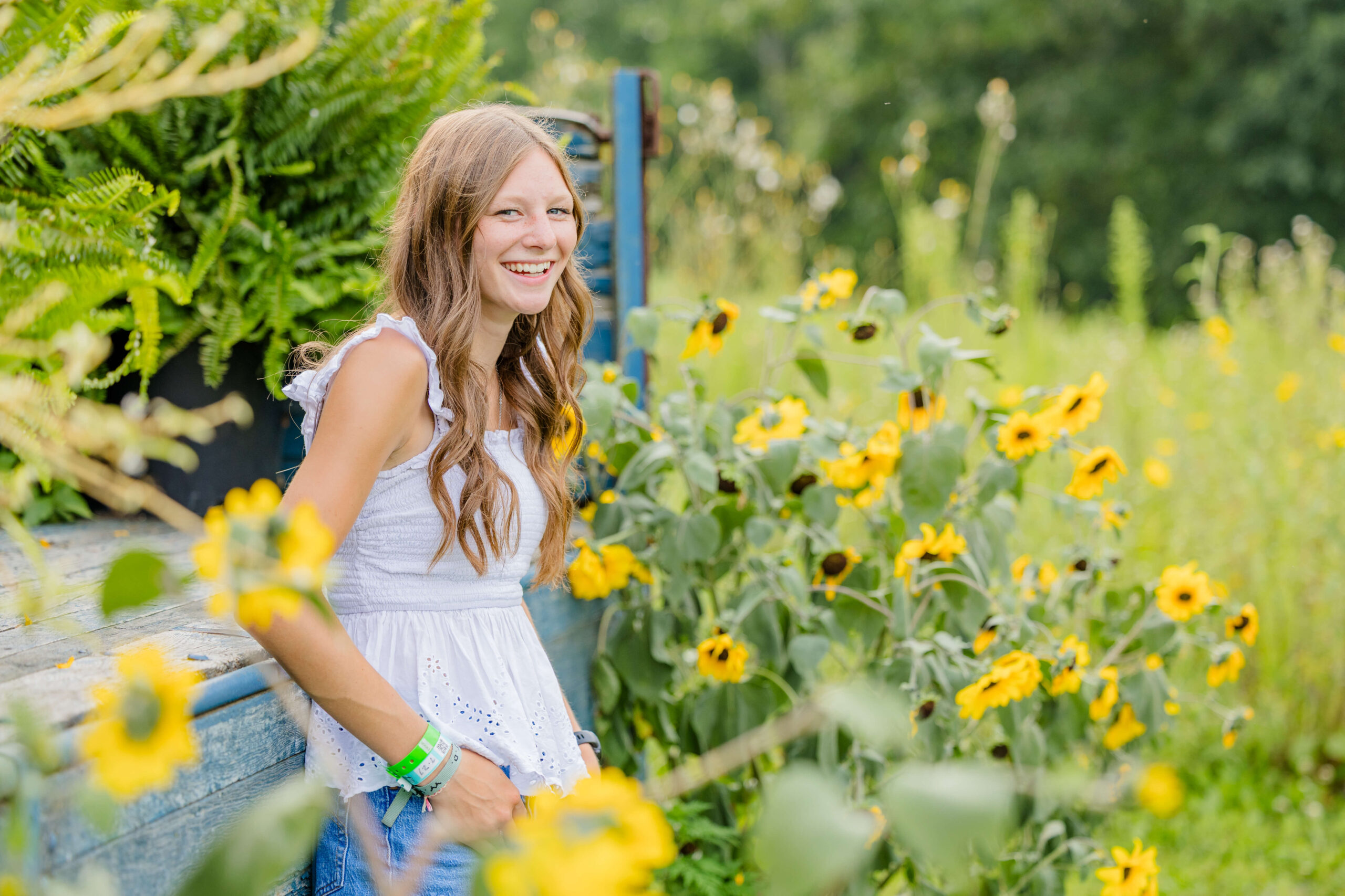 A laughing teenage girl in a white shirt and jeans leans on the back bed of a vintage truck surrounded by flowers