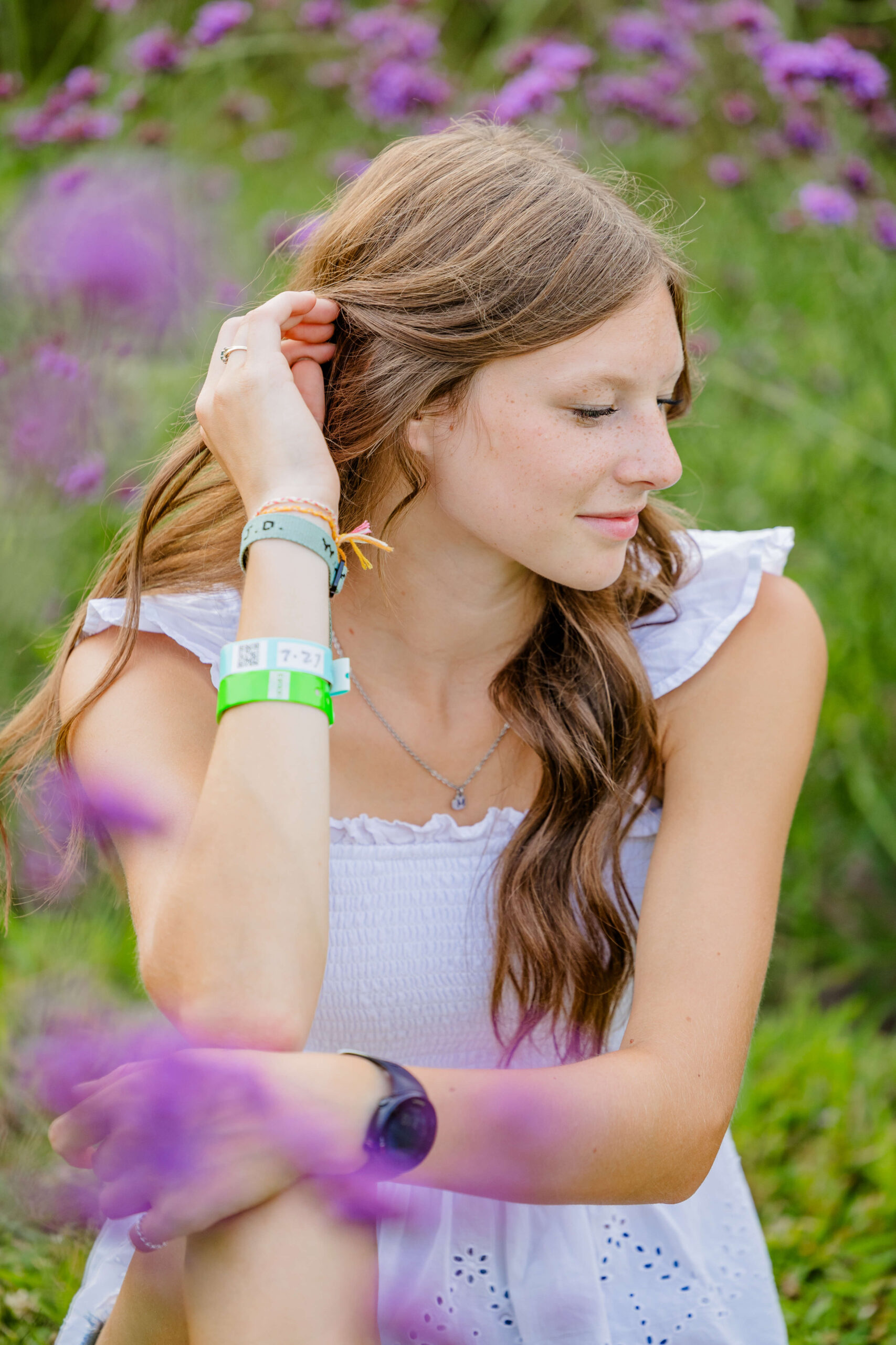 A high school senior in a white dress sits in wildflower fields in michigan pulling her hair from her face
