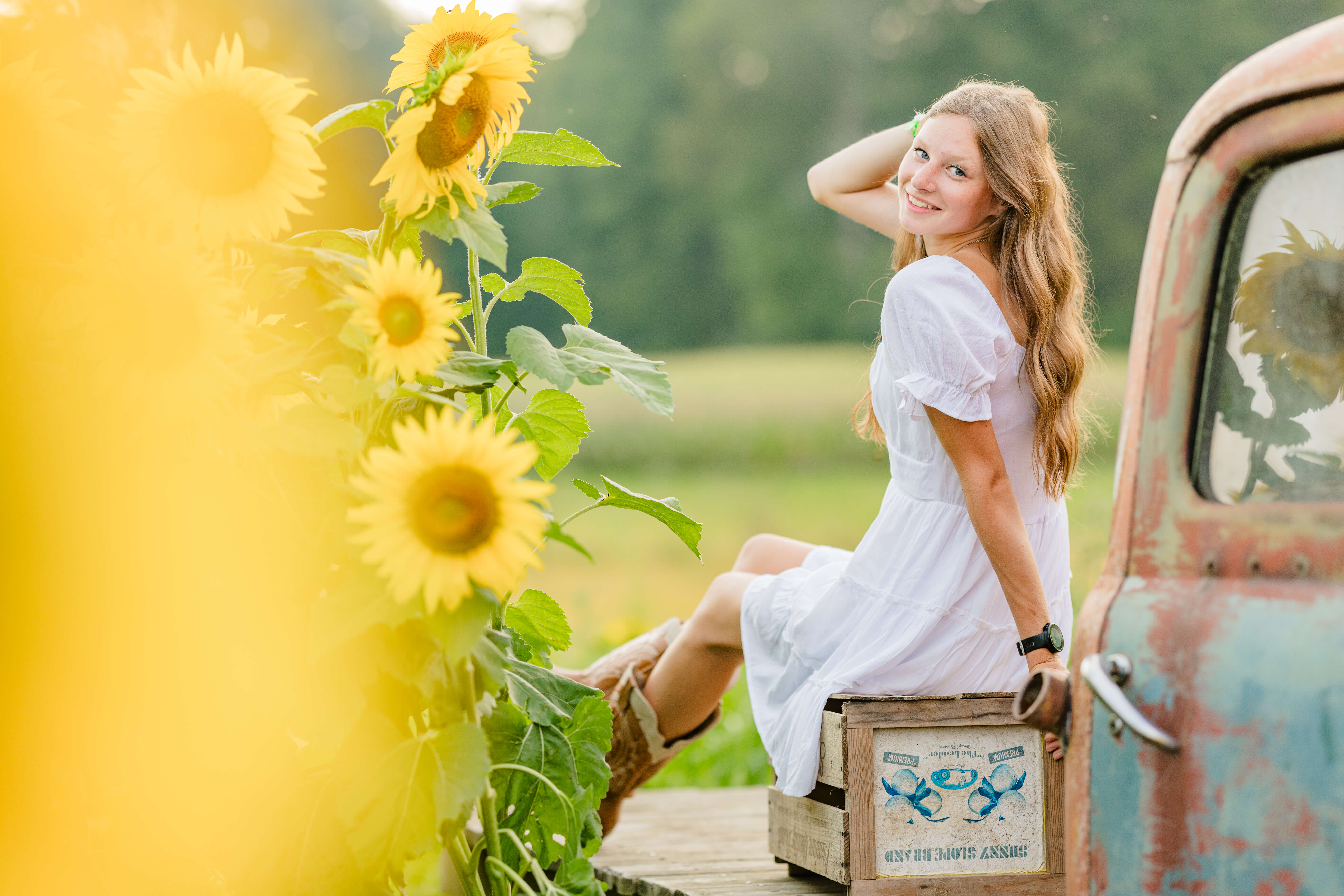 A smiling high school senior in a white dress sits on an apple box on the bed of an antique truck