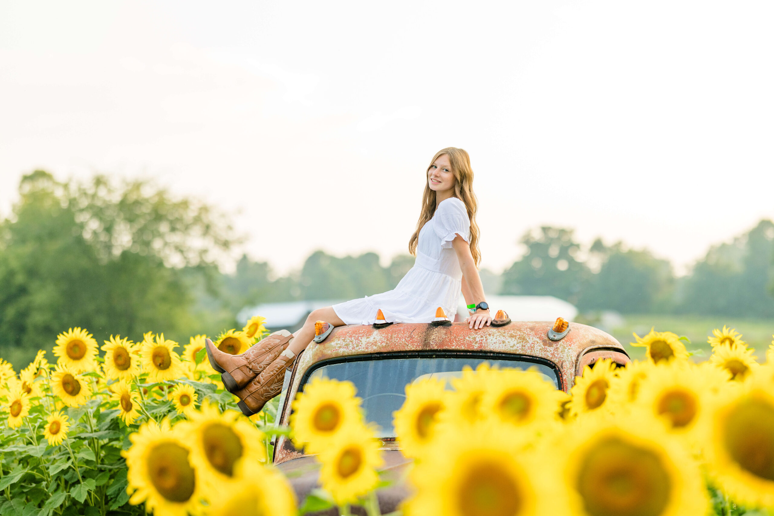 A high school senior in a white dress sits on top of a rusted antique truck overlooking wildflower fields in michigan