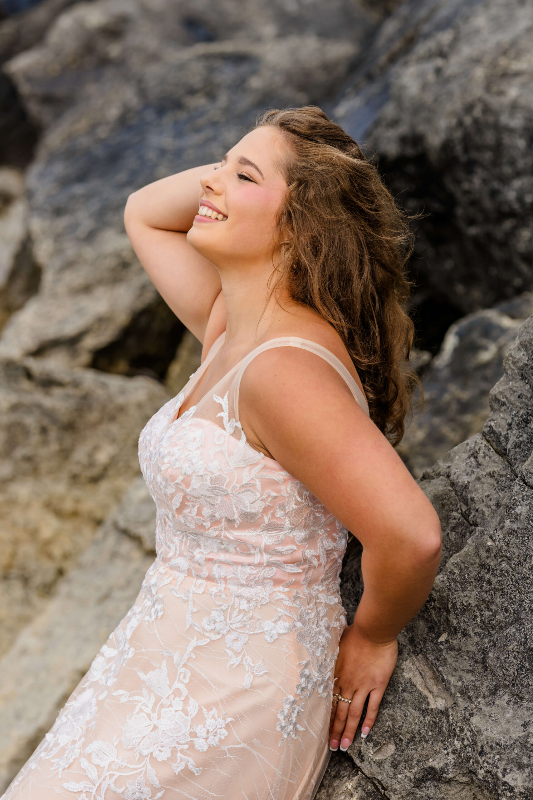 A smiling high school senior leans back on boulders in a pink lace dress with a hand in her hair on one of the beaches near Detroit