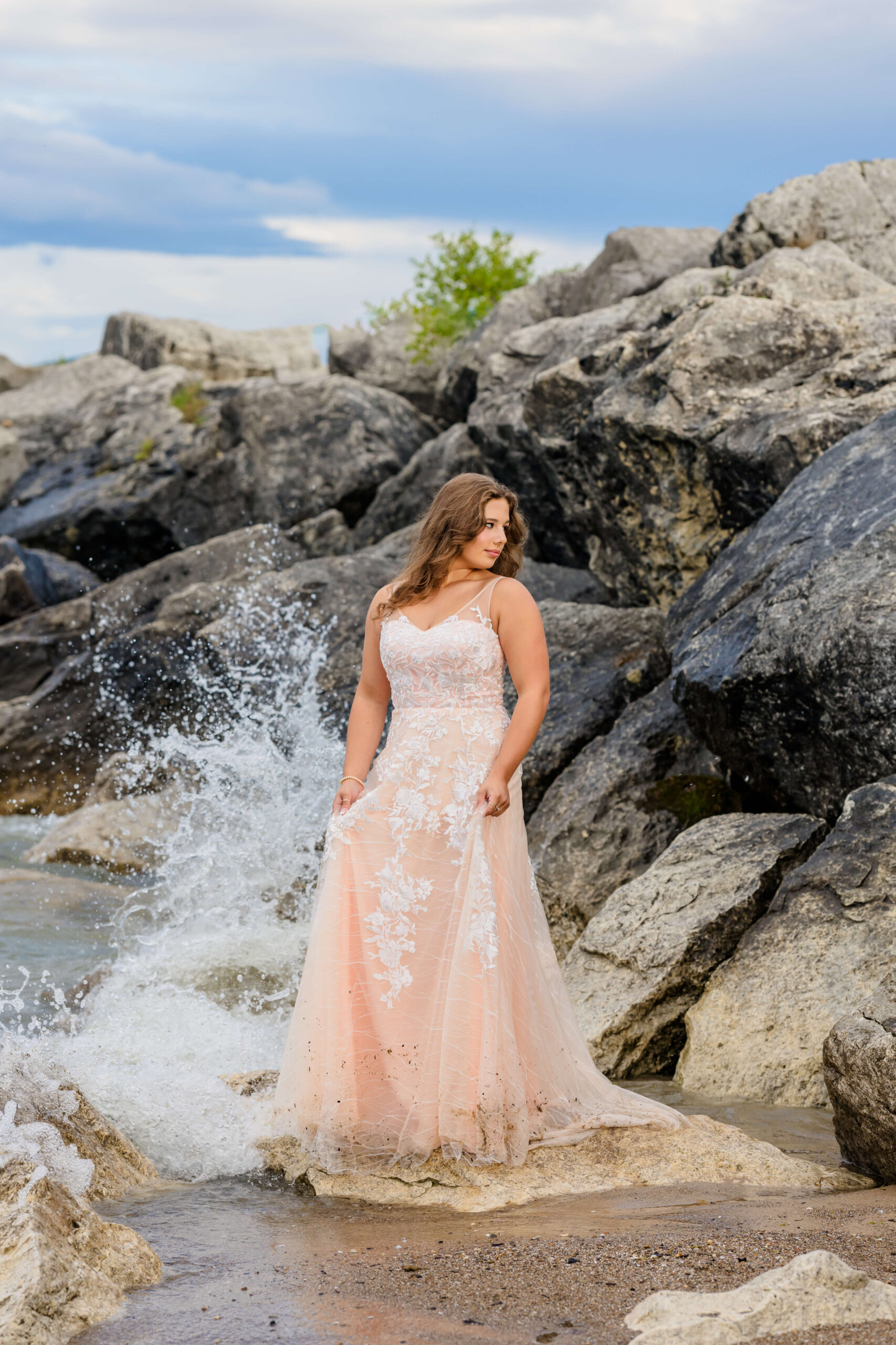A high school senior gazes over her shoulder in a pink lace dress as a wave crashes on a rock while standing on one of the beaches near Detroit