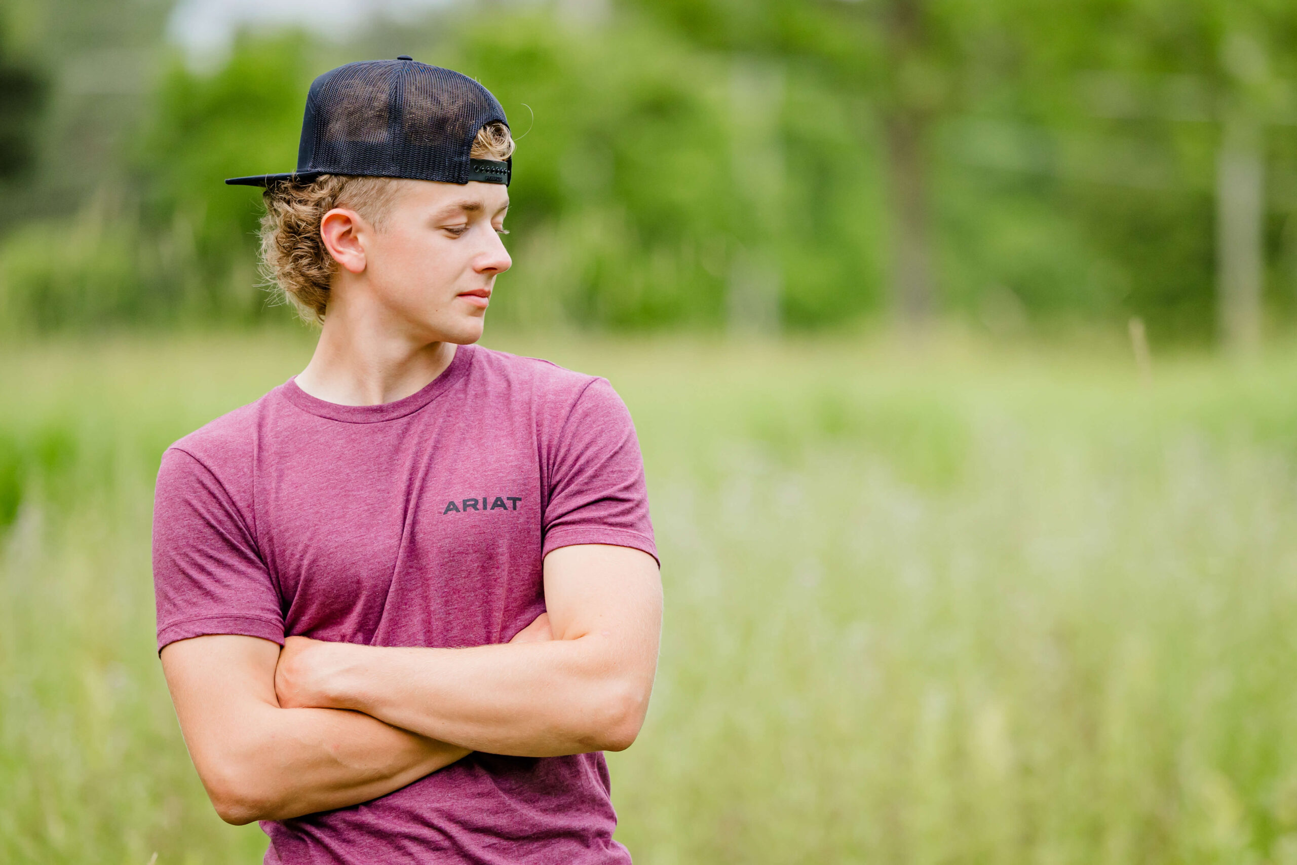 A high school senior stands with arms crossed in a purple shirt and black backwards cap in a field of tall grass at one of the parks near detroit