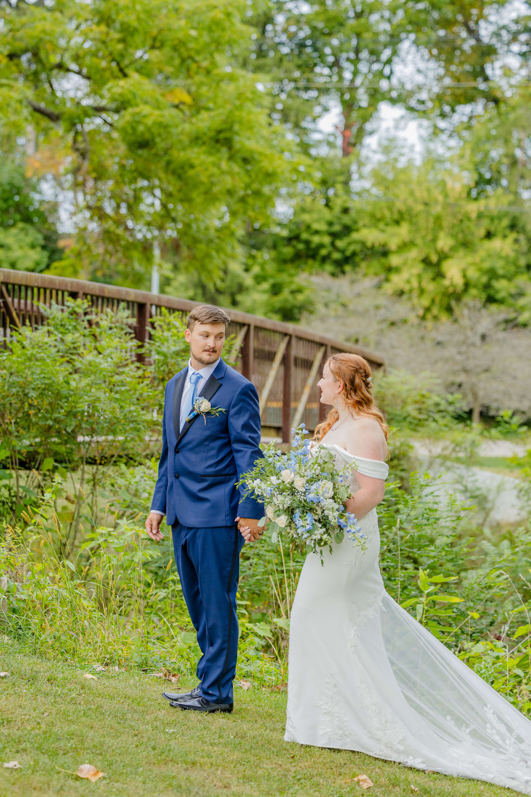A groom walks with his bride looking at each other along the water by a pedestrian bridge