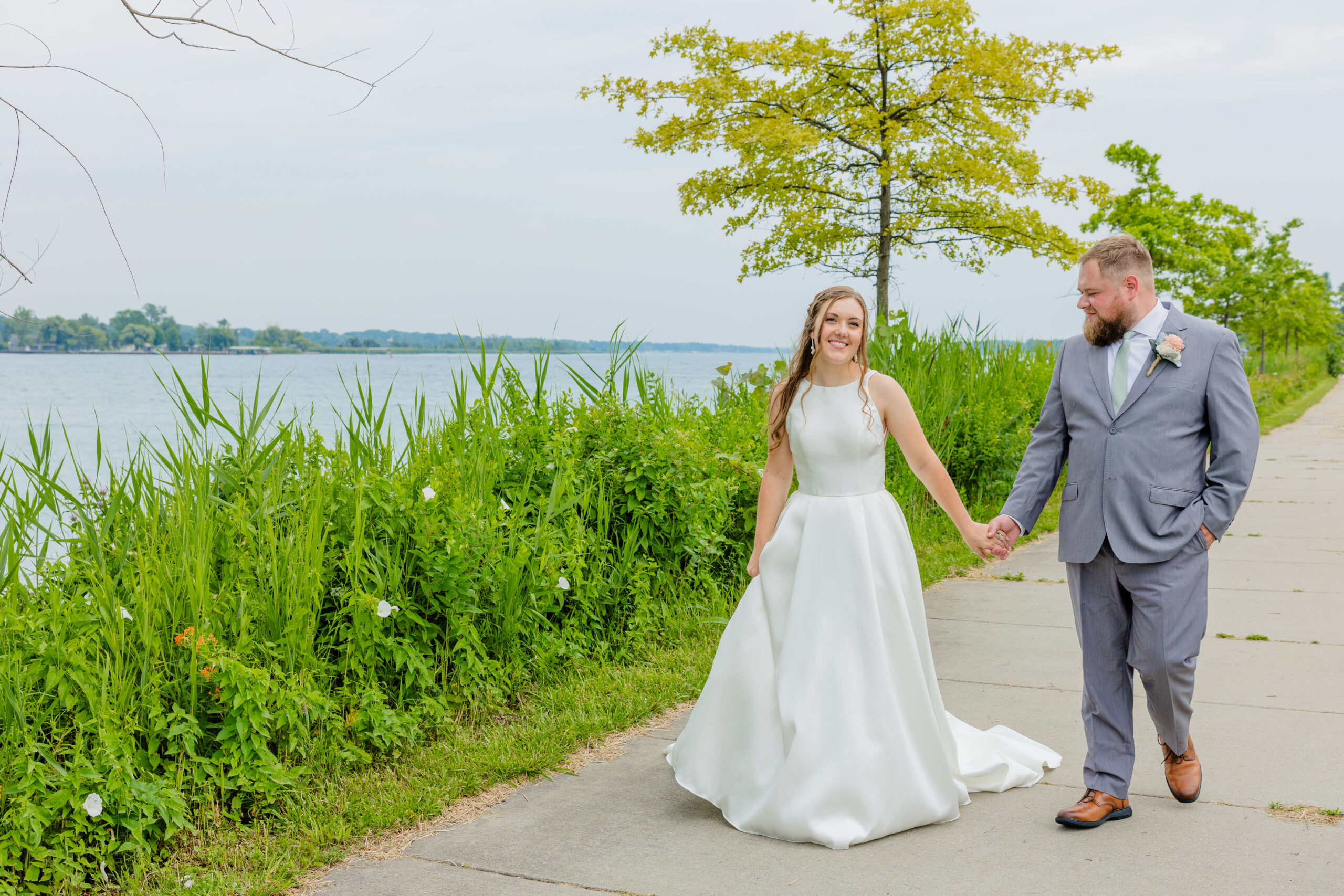 A bride walks with her groom on the sidewalk holding hands and smiling by the water