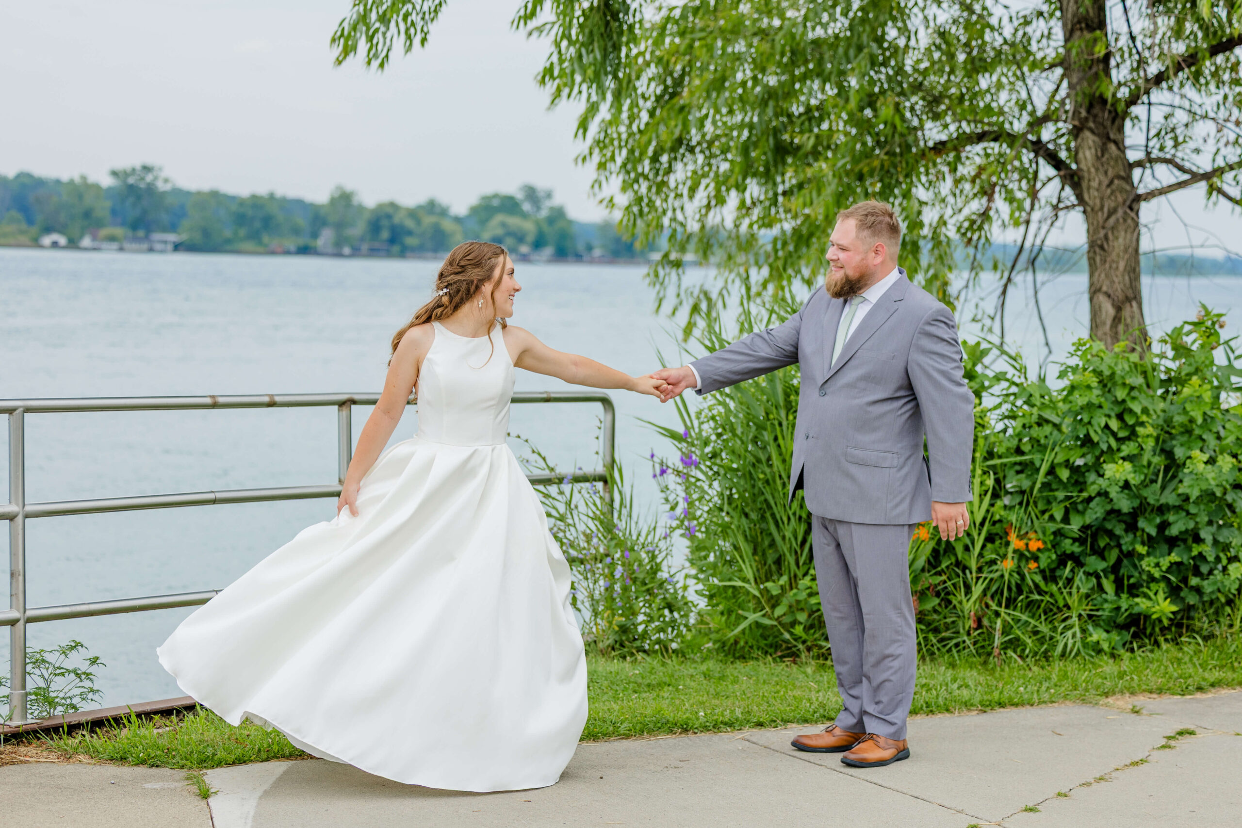A bride leads her groom by the hand on a waterfront walk during their alexanders banquet hall wedding