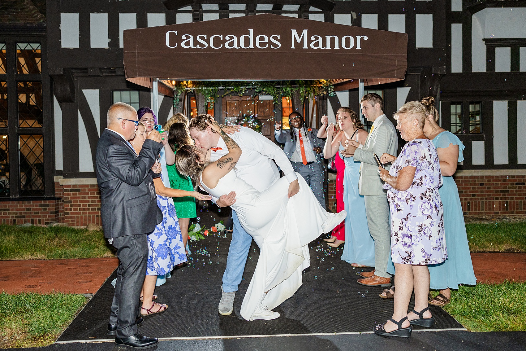 Newlyweds in white kiss with a dip under bubbles blown by happy guests while exiting the cascades manor wedding venue