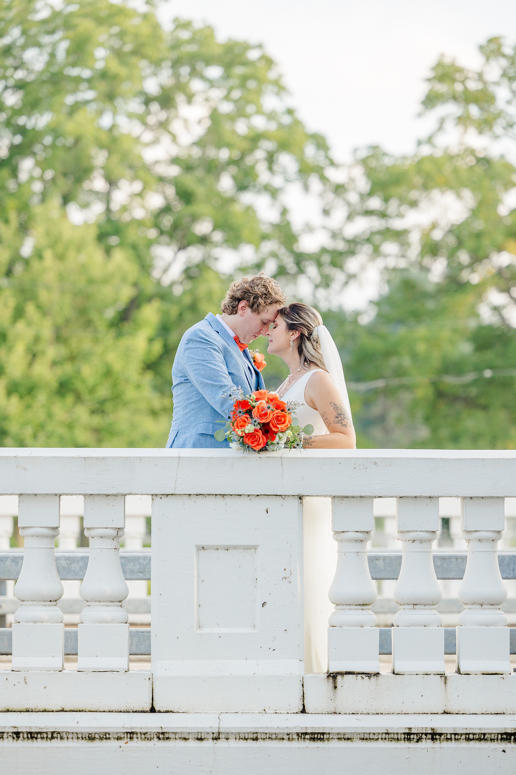 A bride and groom nuzzle foreheads while standing on a white bridge at sunset