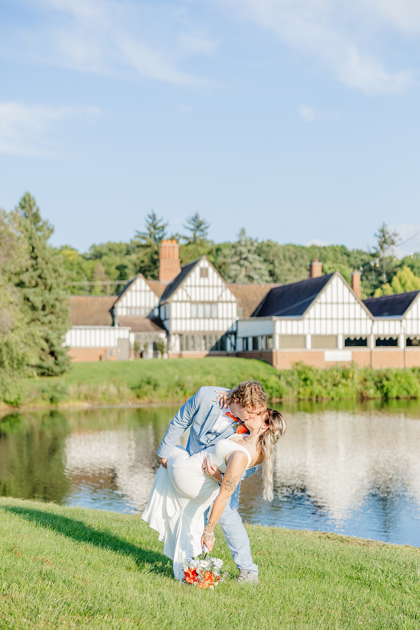 A groom in blue dips his bride for a kiss by a pond
