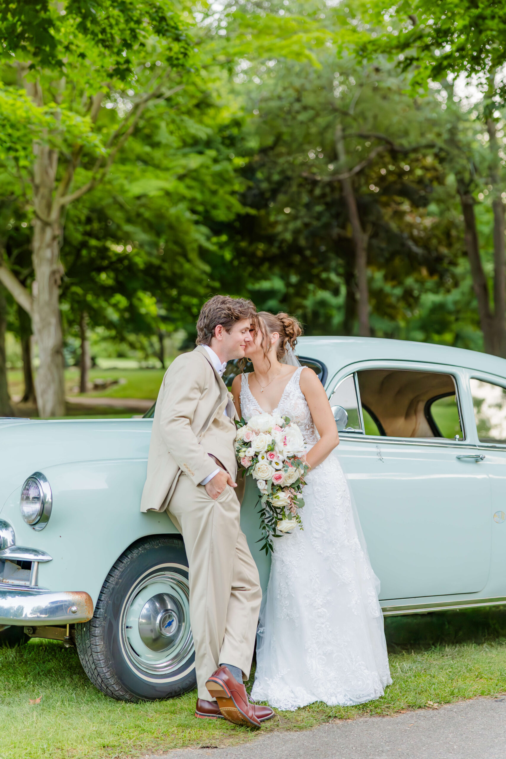 A bride kisses her groom's cheek as they lean against a vintage car during their golden hawk golf course wedding