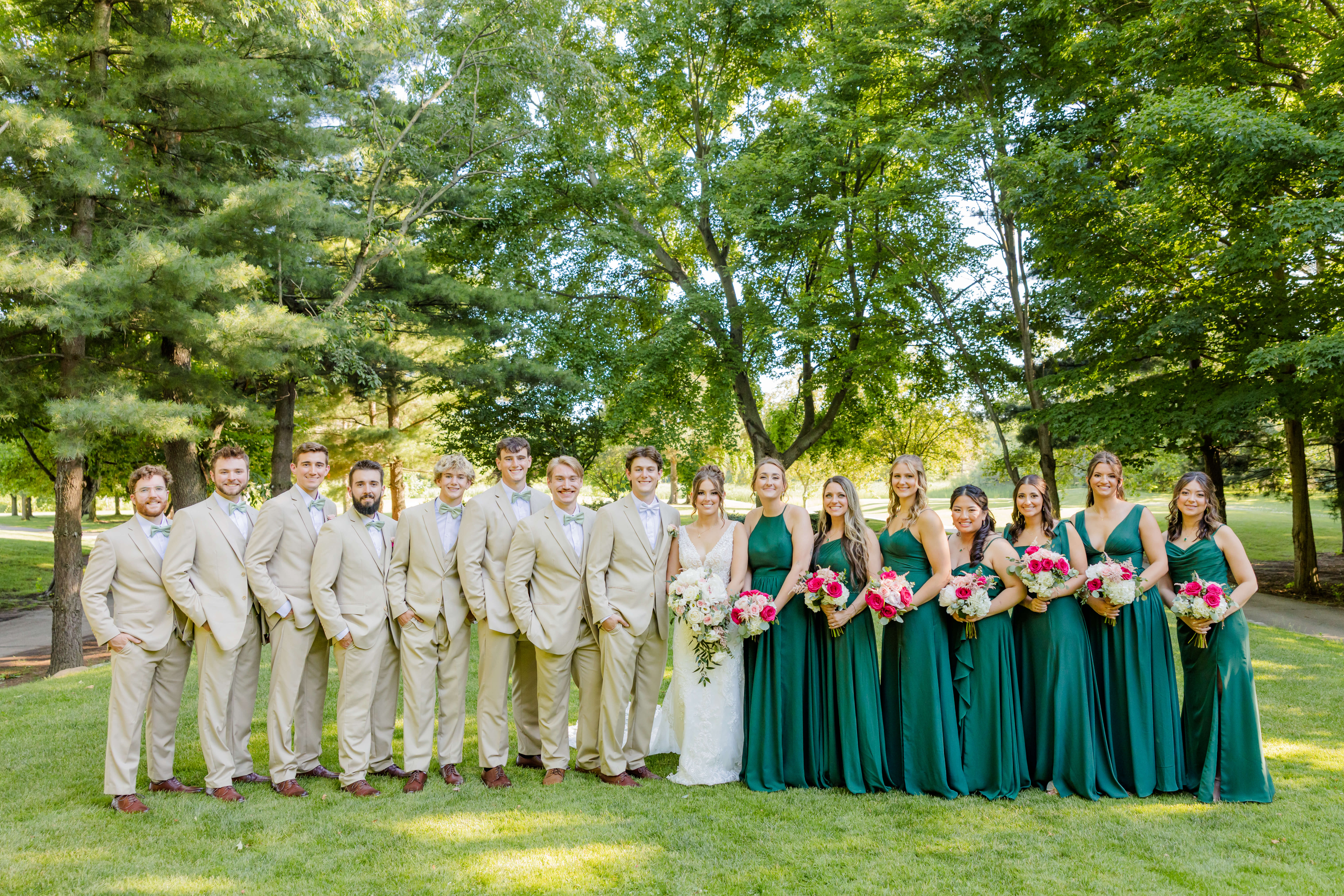 A bride and groom stand with their large weeding party in green and tan in the lawn