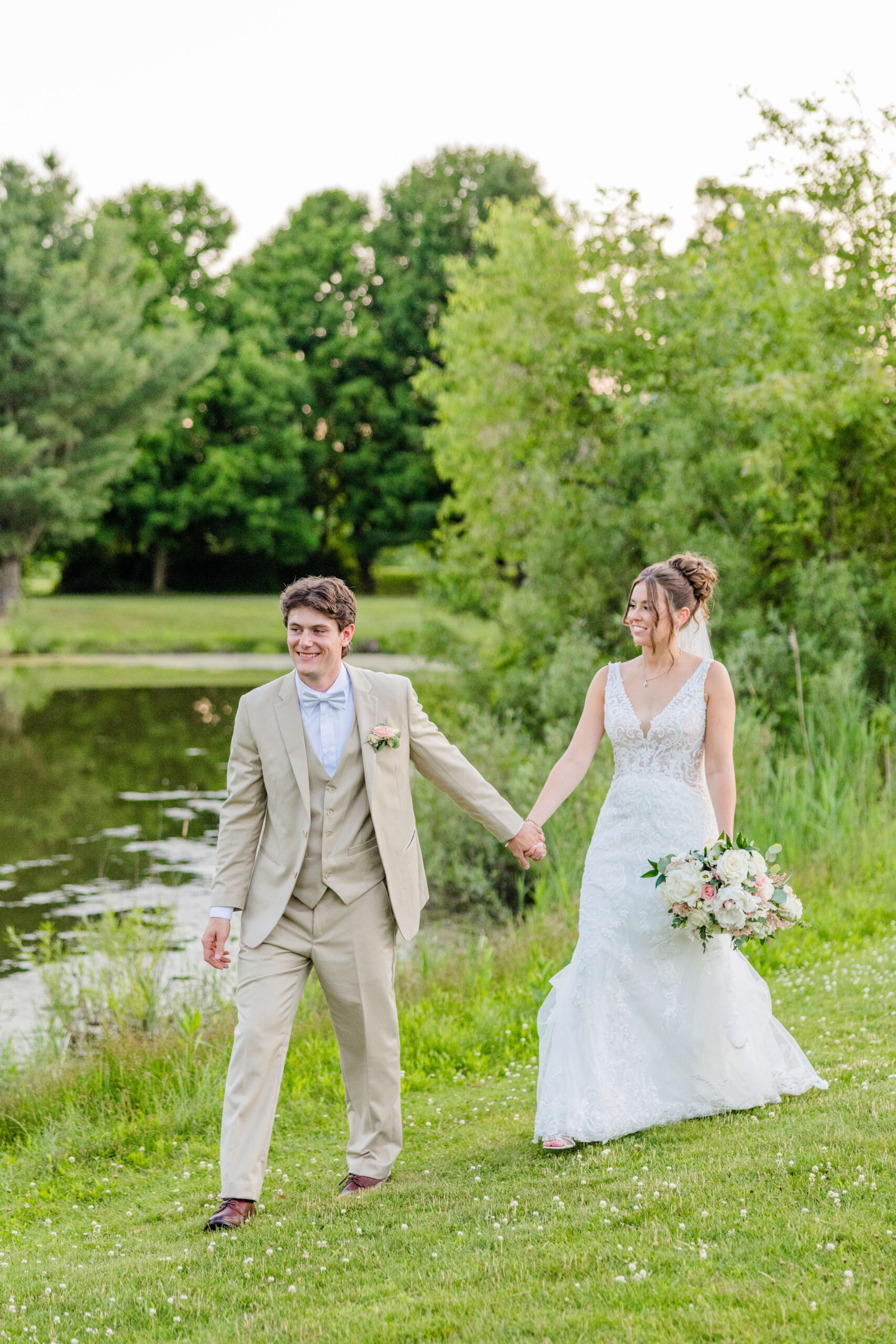 A groom in a tan suit leads his bride on a sunset walk around a pond