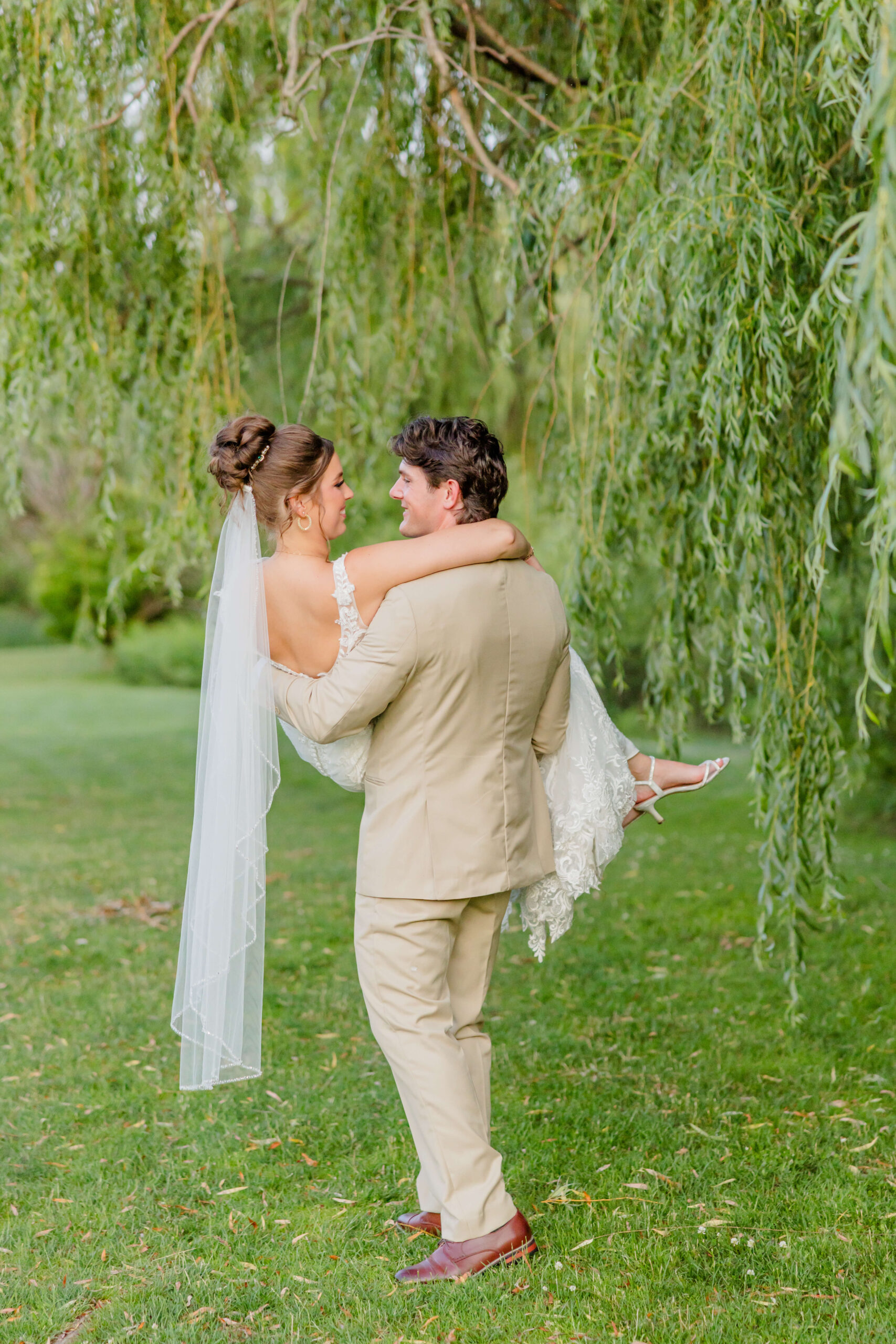 A groom in a tan suit carries his bride under a willow tree in a lawn