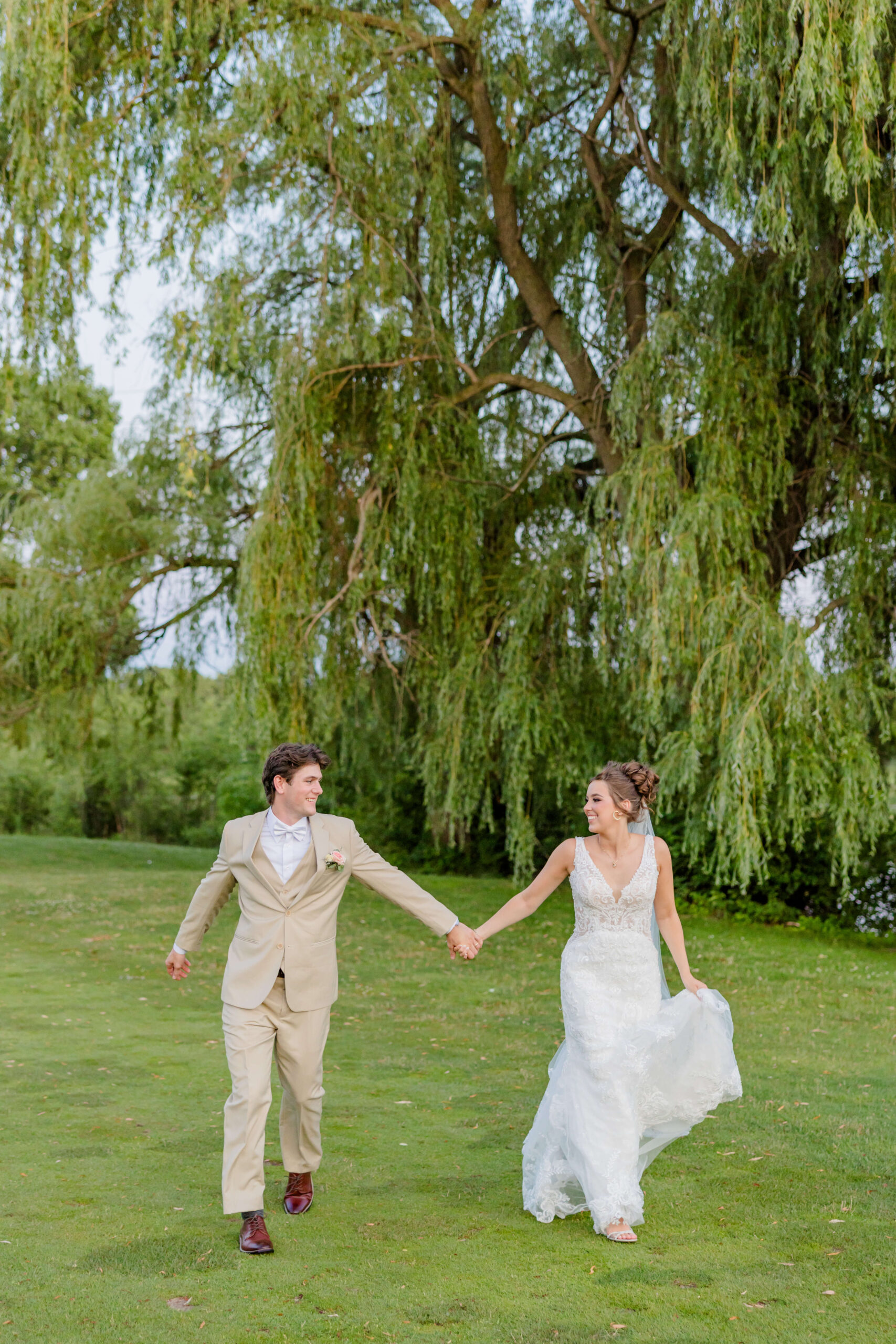 Newlyweds happily walk hand in hand under a willow tree in a lawn during their golden hawk golf course wedding