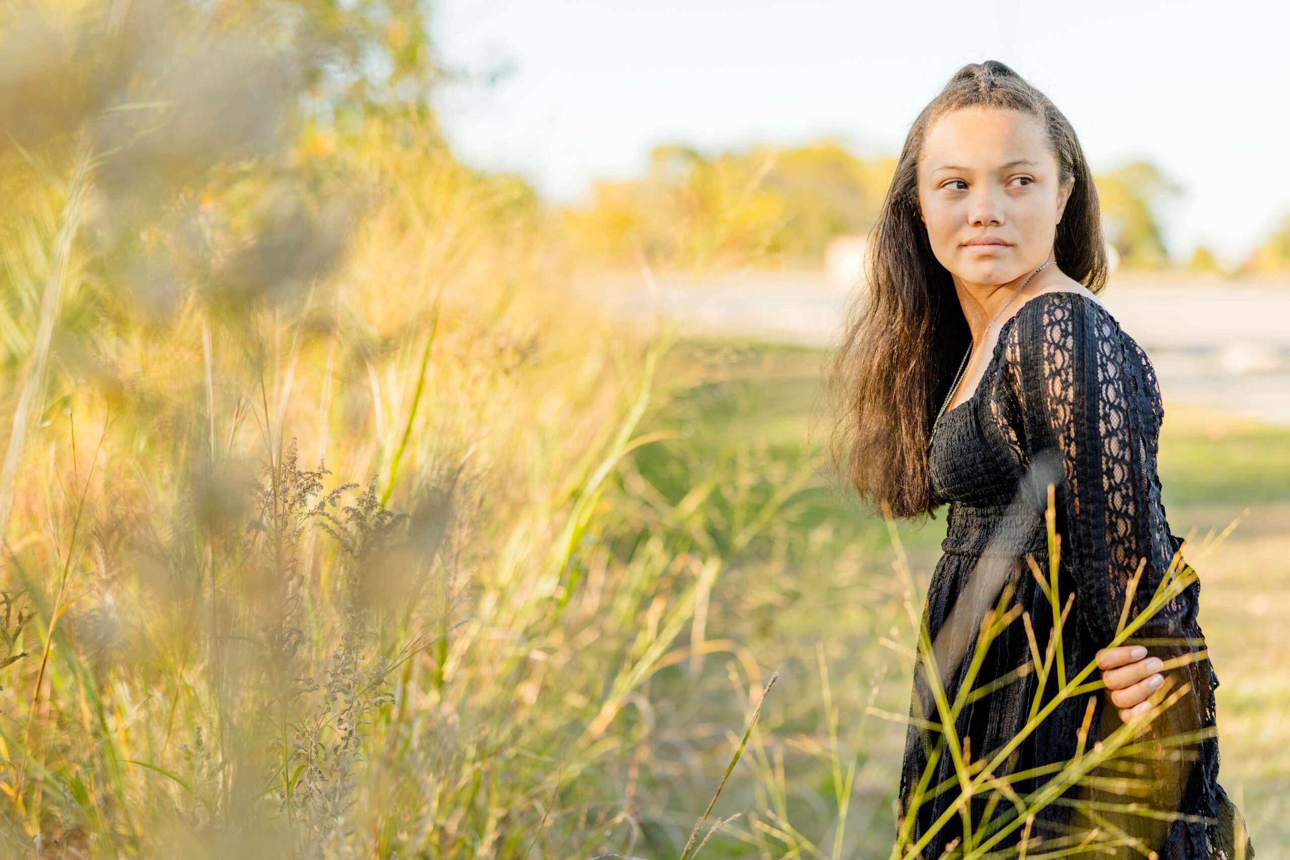 A high school senior in a black dress with long dark hair walks in tall golden grass while looking back over her shoulder