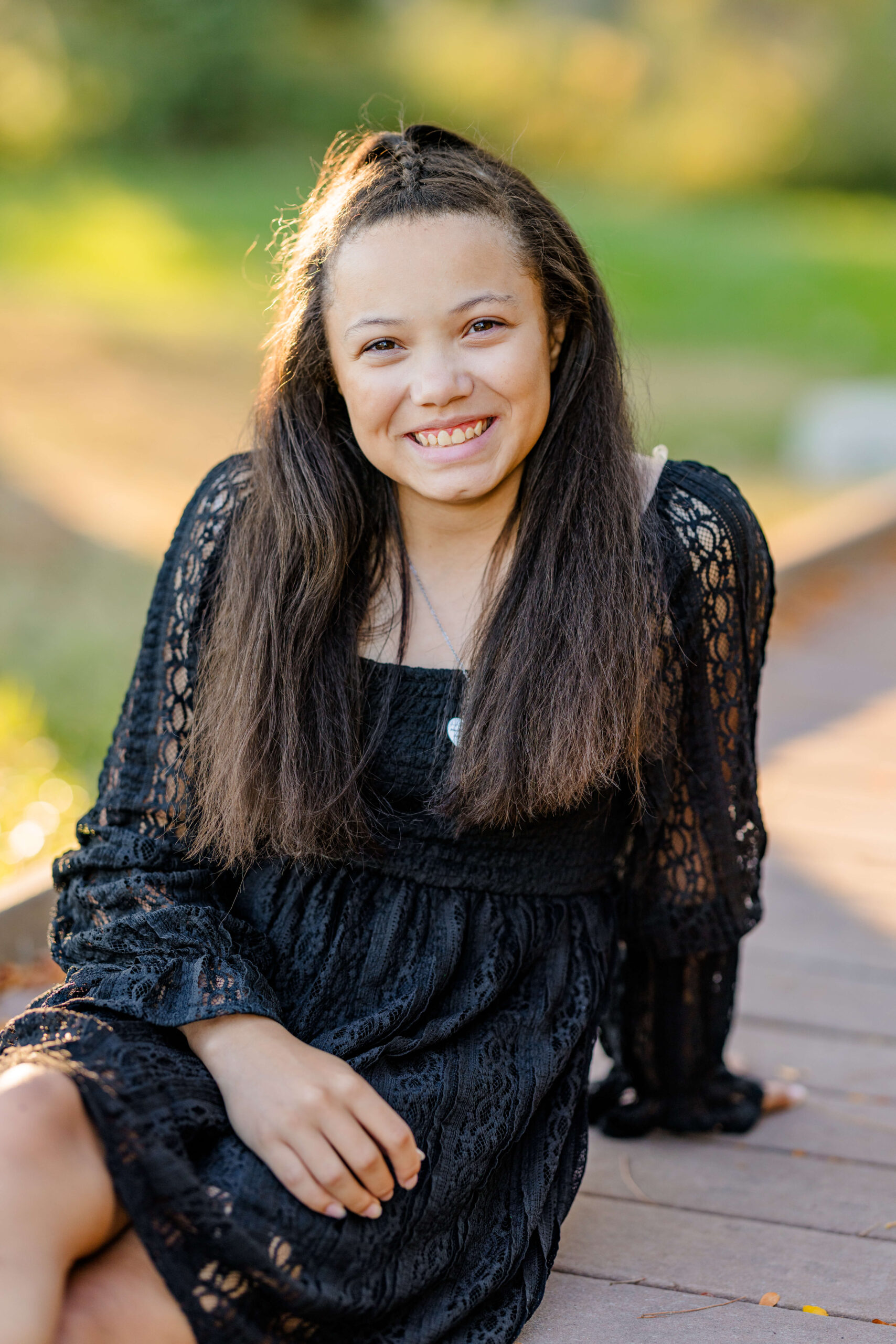 A smiling high school senior in a black lace dress sits on a dock at sunset after finding beautiful prom dresses in michigan