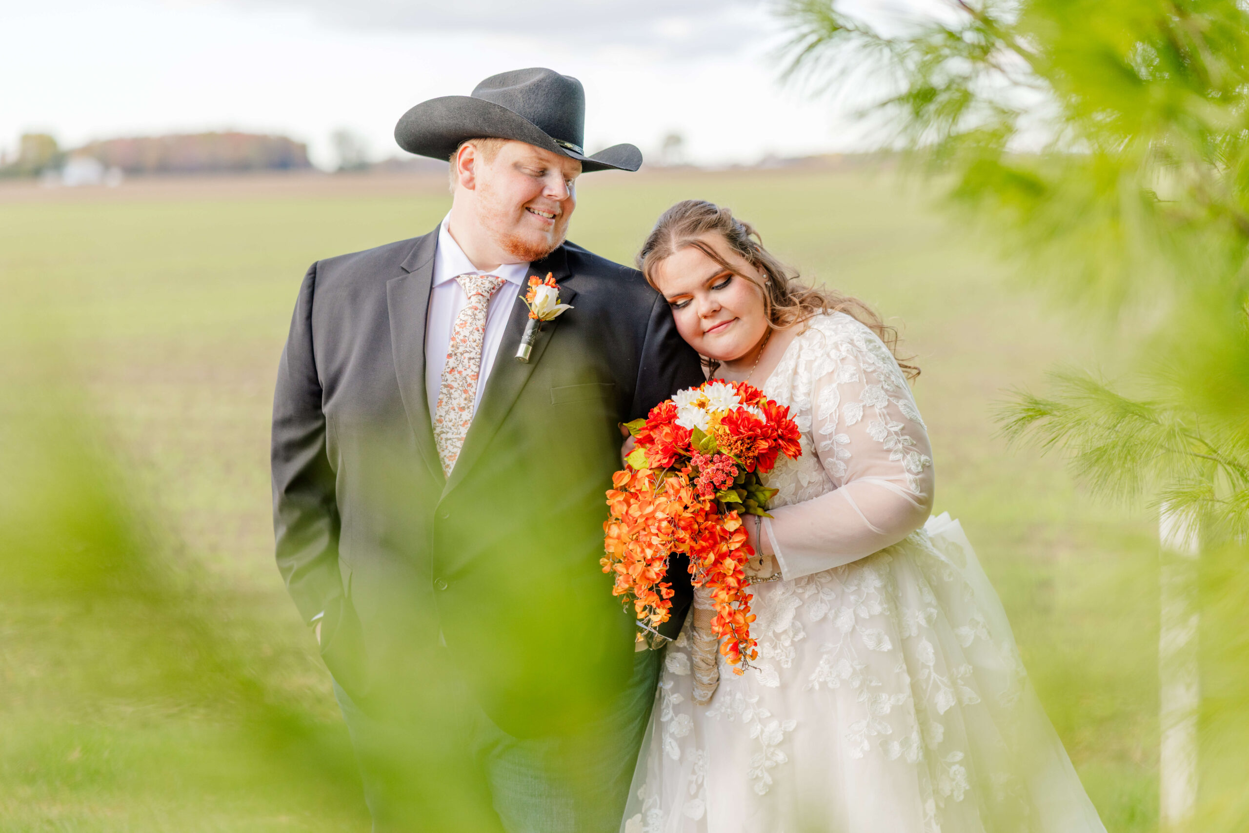 A bride admires her bright red bouquet in her hand while leaning on her groom's shoulder