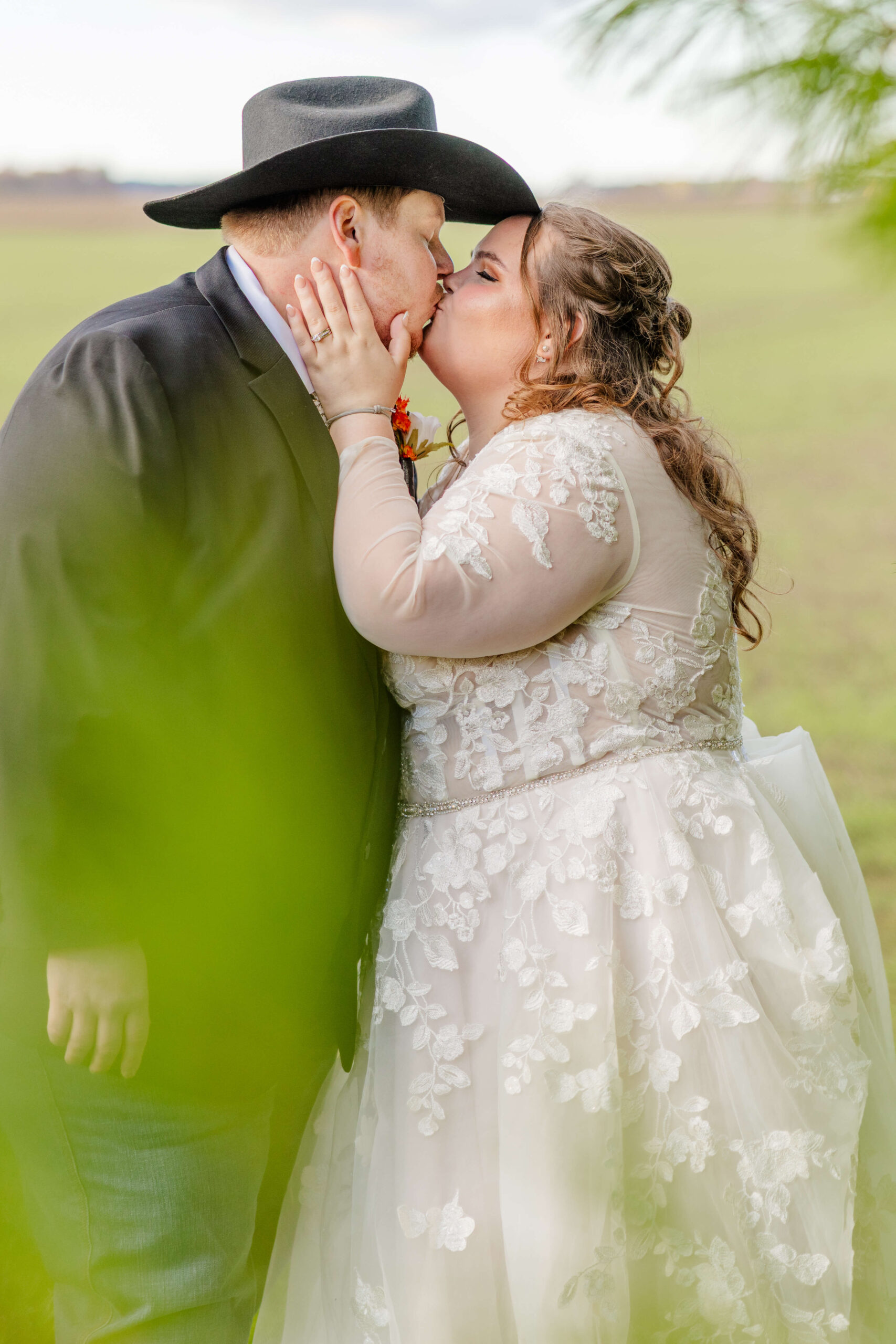 A bride holds and kisses her groom under his cowboy hat in a pasture during their sutton barn wedding