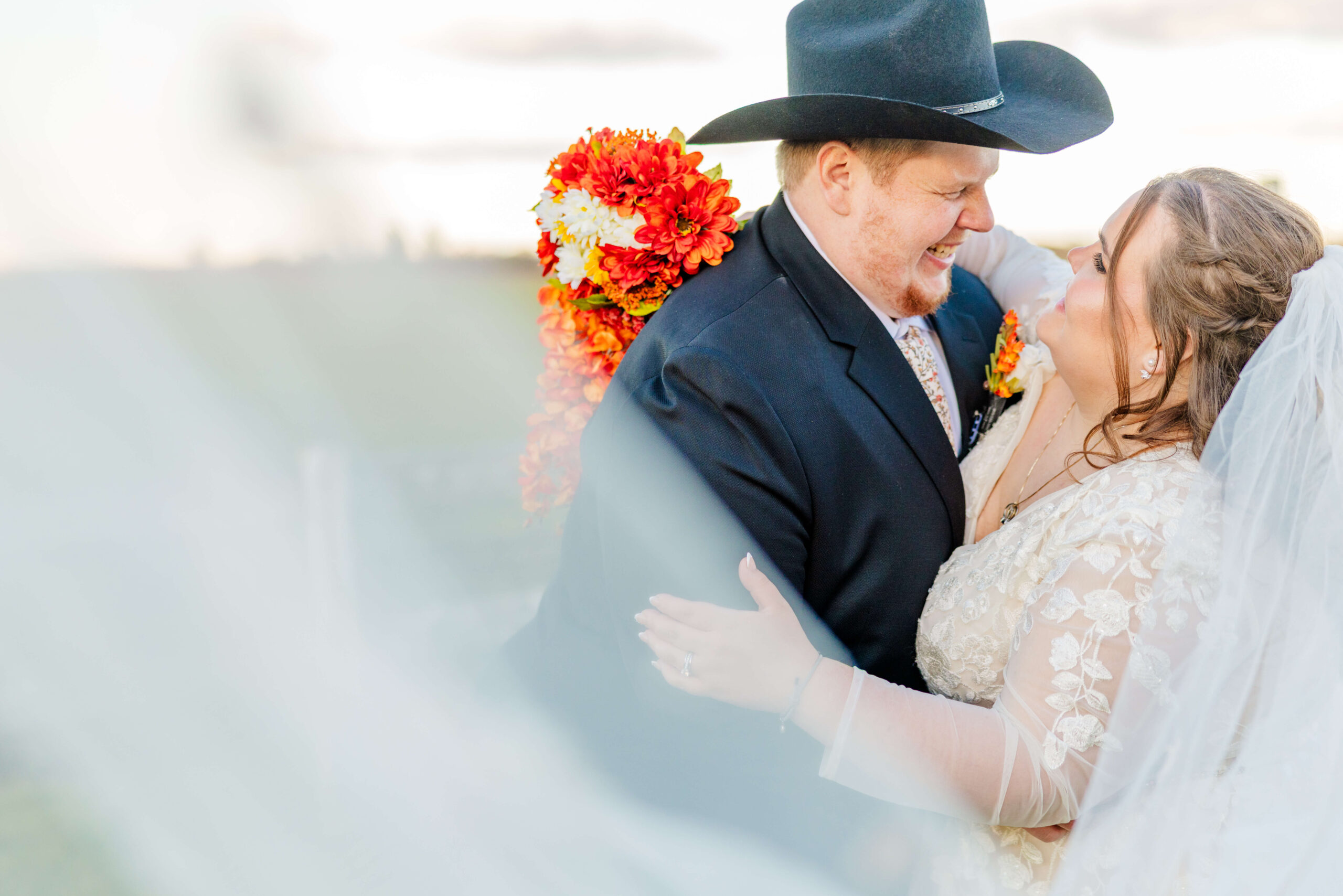 A happy couple dance and laugh in a black suit and cowboy hat and lace embroidered dress in a pasture at sunset during their sutton barn wedding