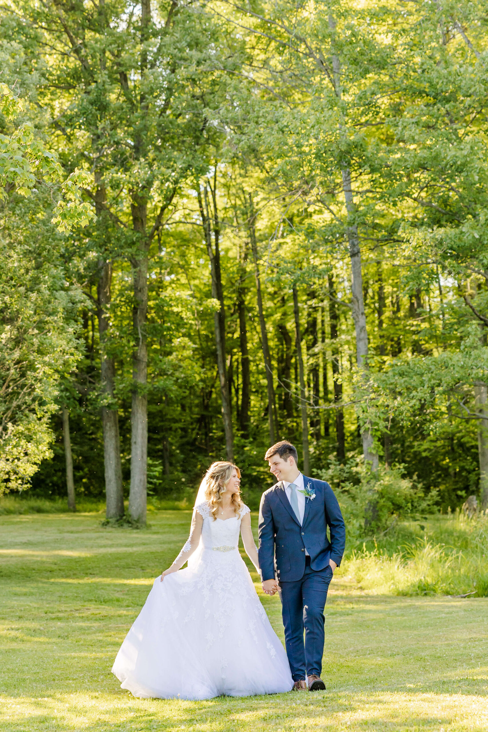 A bride and groom laugh while walking holding hands out of a forest