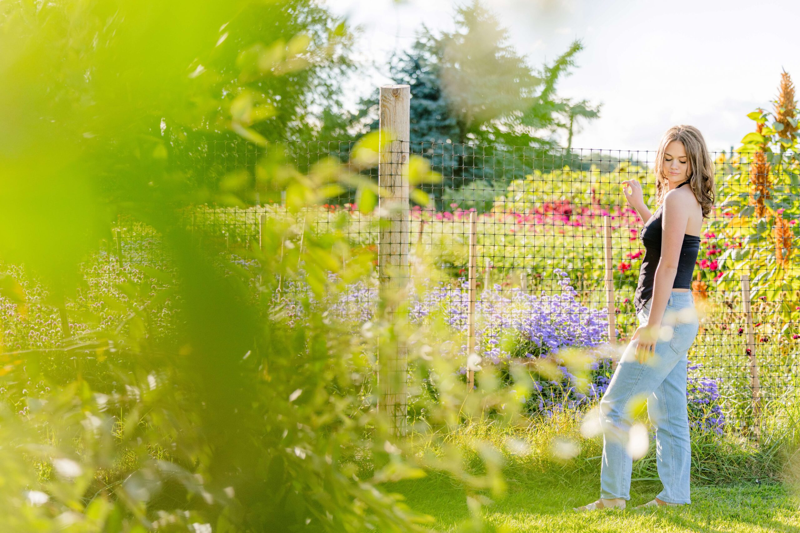 A high school senior in a black blouse and jeans explores a flower garden at sunset