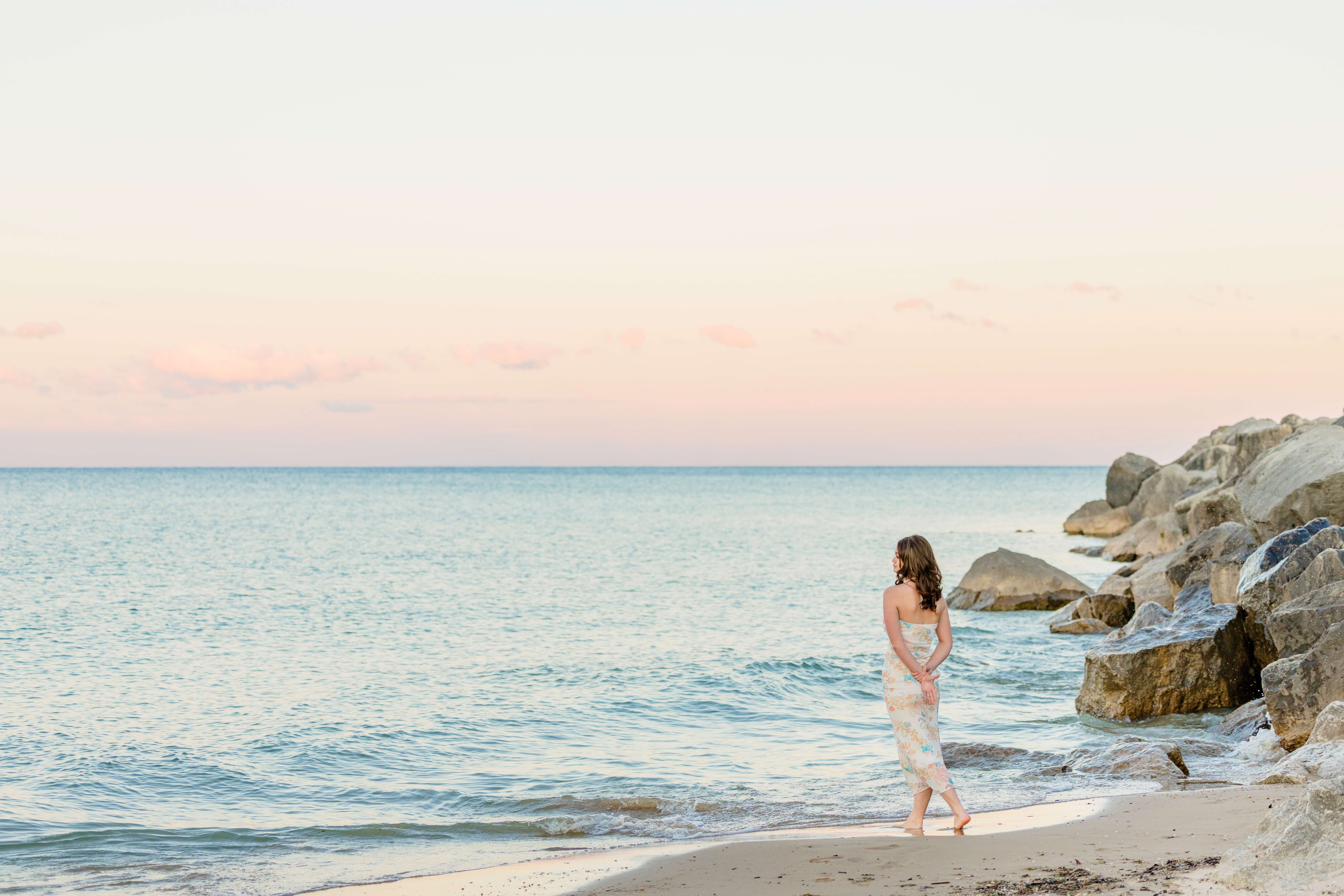 A high school senior walks on the beach by the water in a flower print dress by a jetty after finding hair salons in port huron at sunset