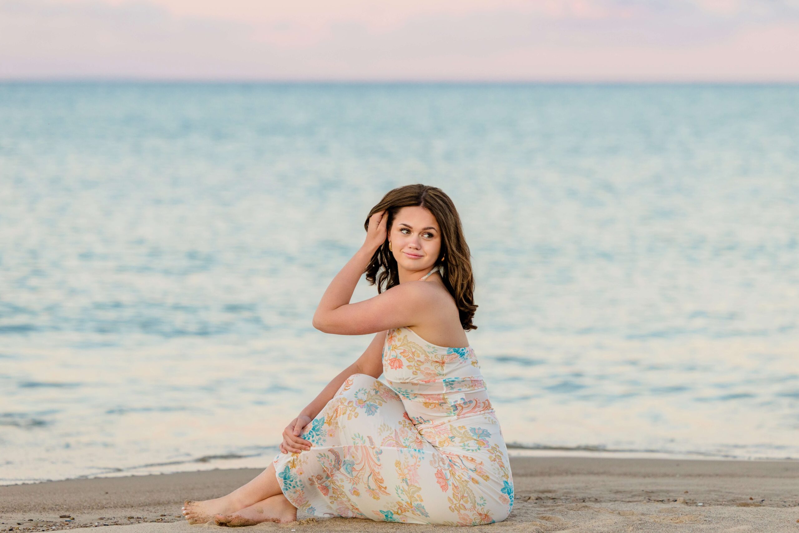 A high school senior in a flower print dress sits in the sand on the beach pulling her hair back at sunset
