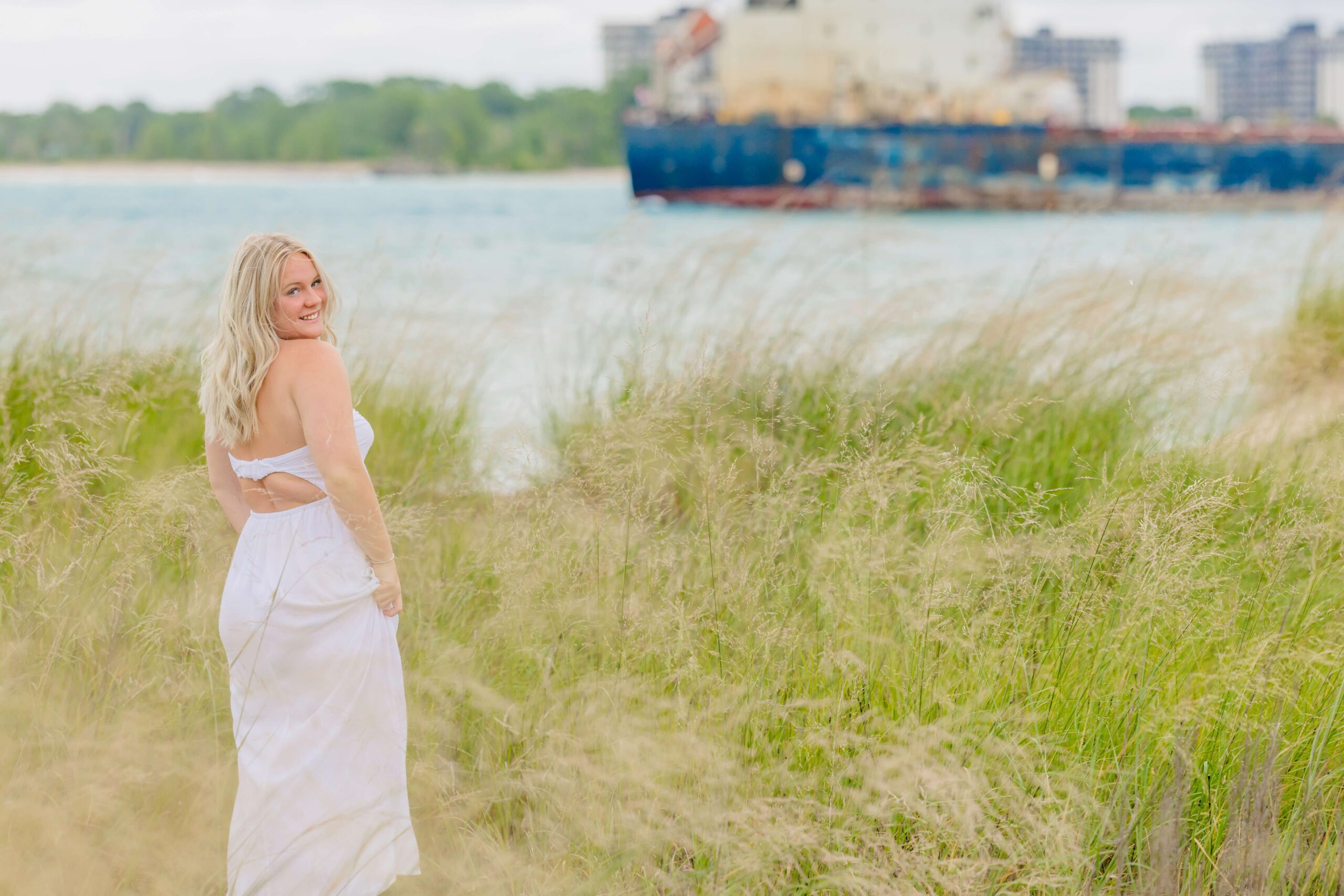 A woman in a white dress smiles over her shoulder while walking in tall grass towards the water