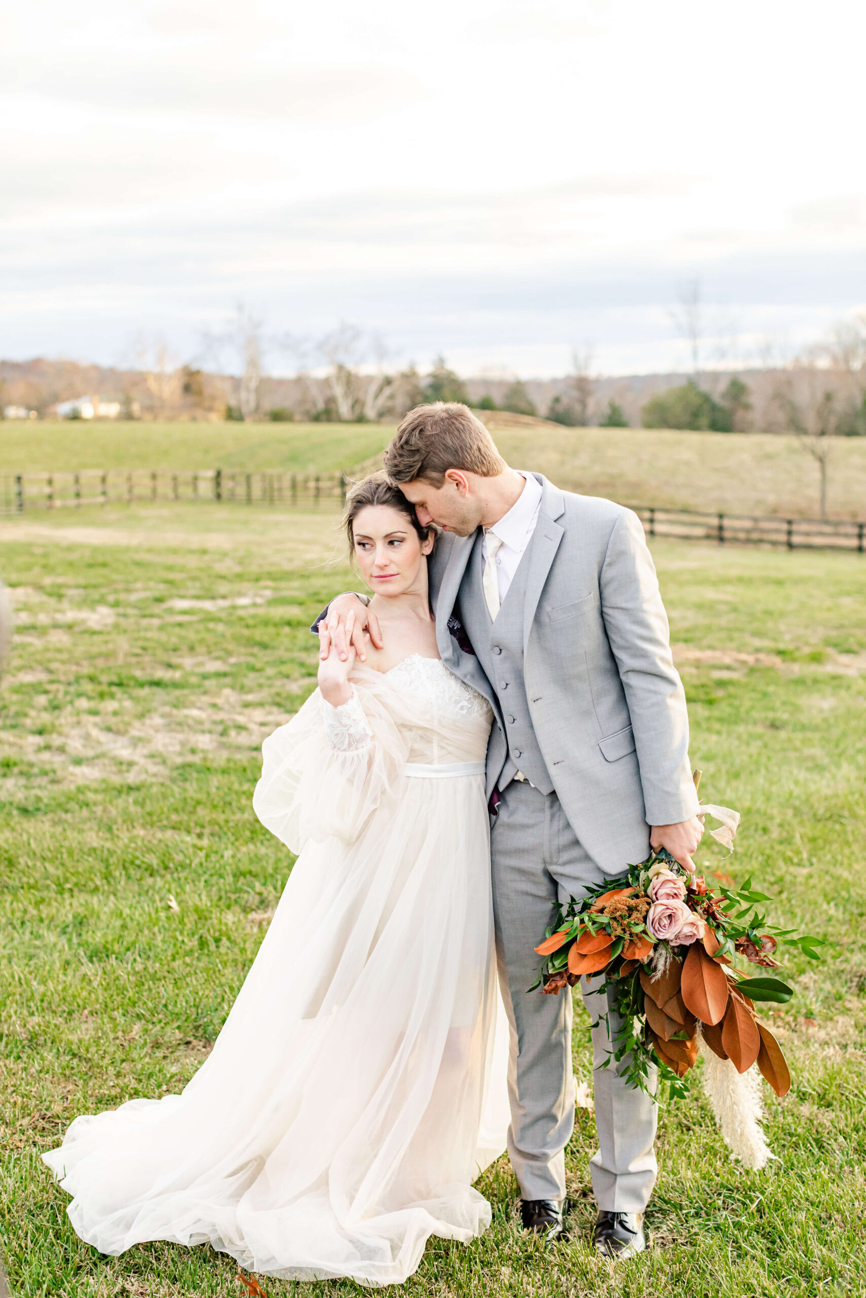 Newlyweds hold hands on the bride's shoulder while snuggling in a pasture at sunset at one of the rustic wedding venues in michigan