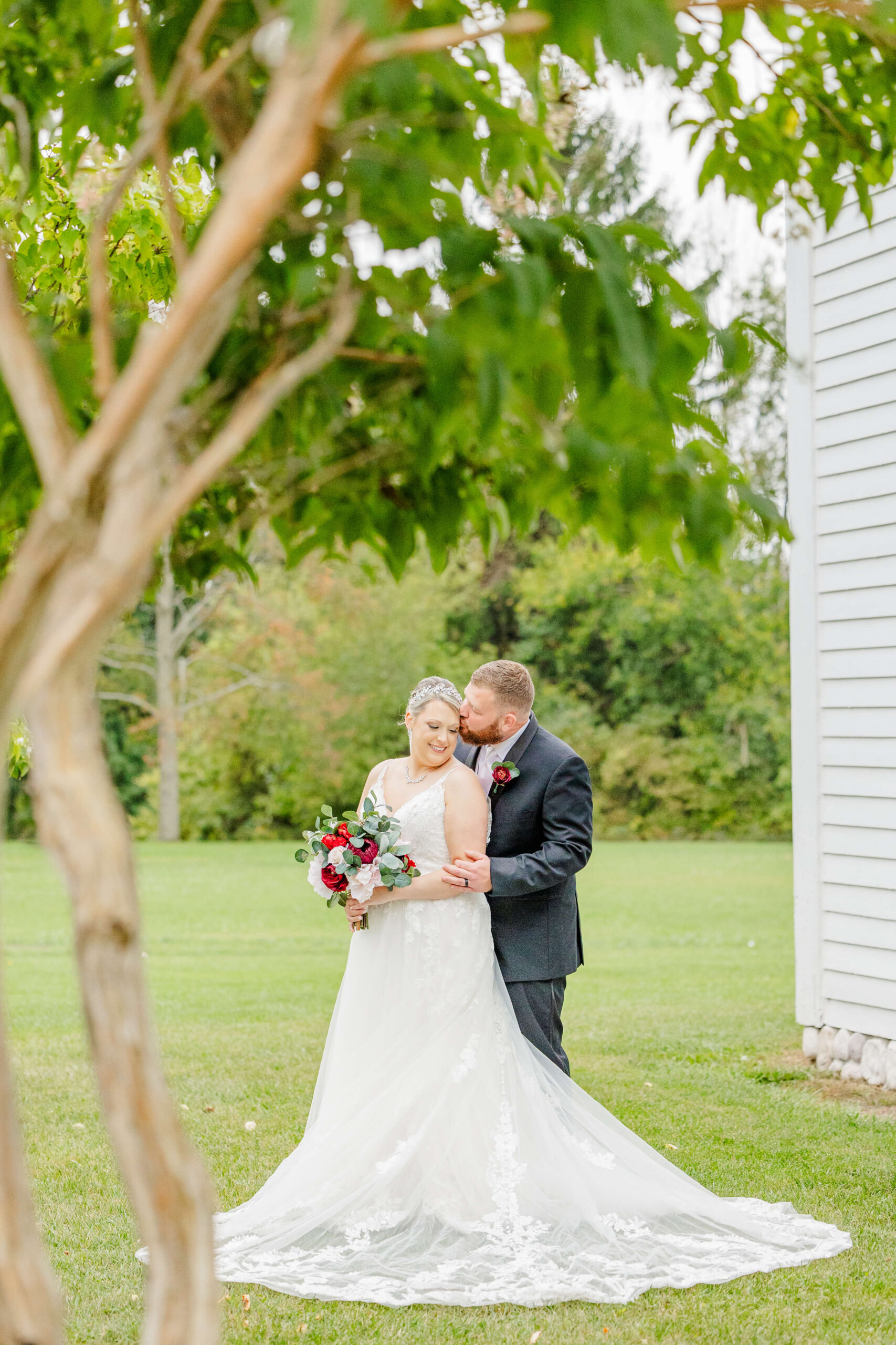 A groom kisses the temple of his smiling bride as they snuggle in the lawn under green trees