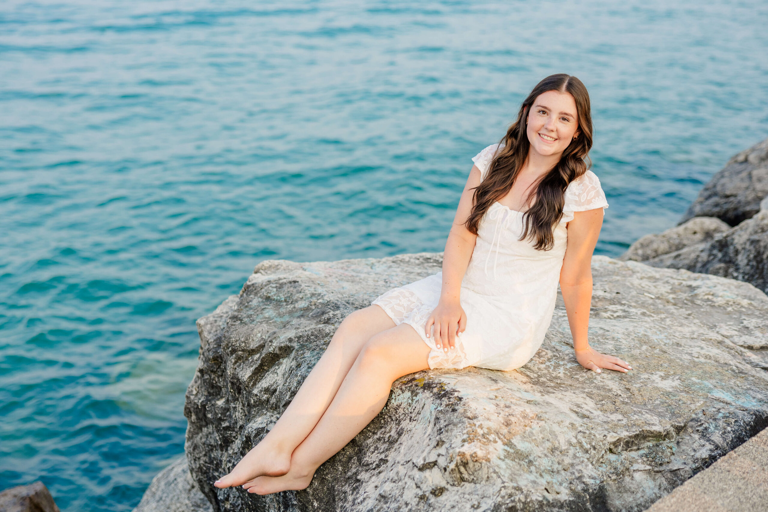 A woman smiles while sitting on a boulder overlooking the blue water in a white dress after finding everything for prom in port huron