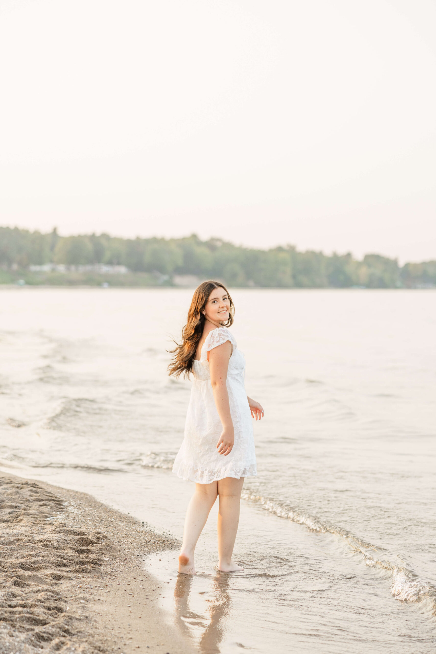 A happy high school senior in a white dress looks over her shoulder while walking on a beach at sunset before heading to prom in port huron