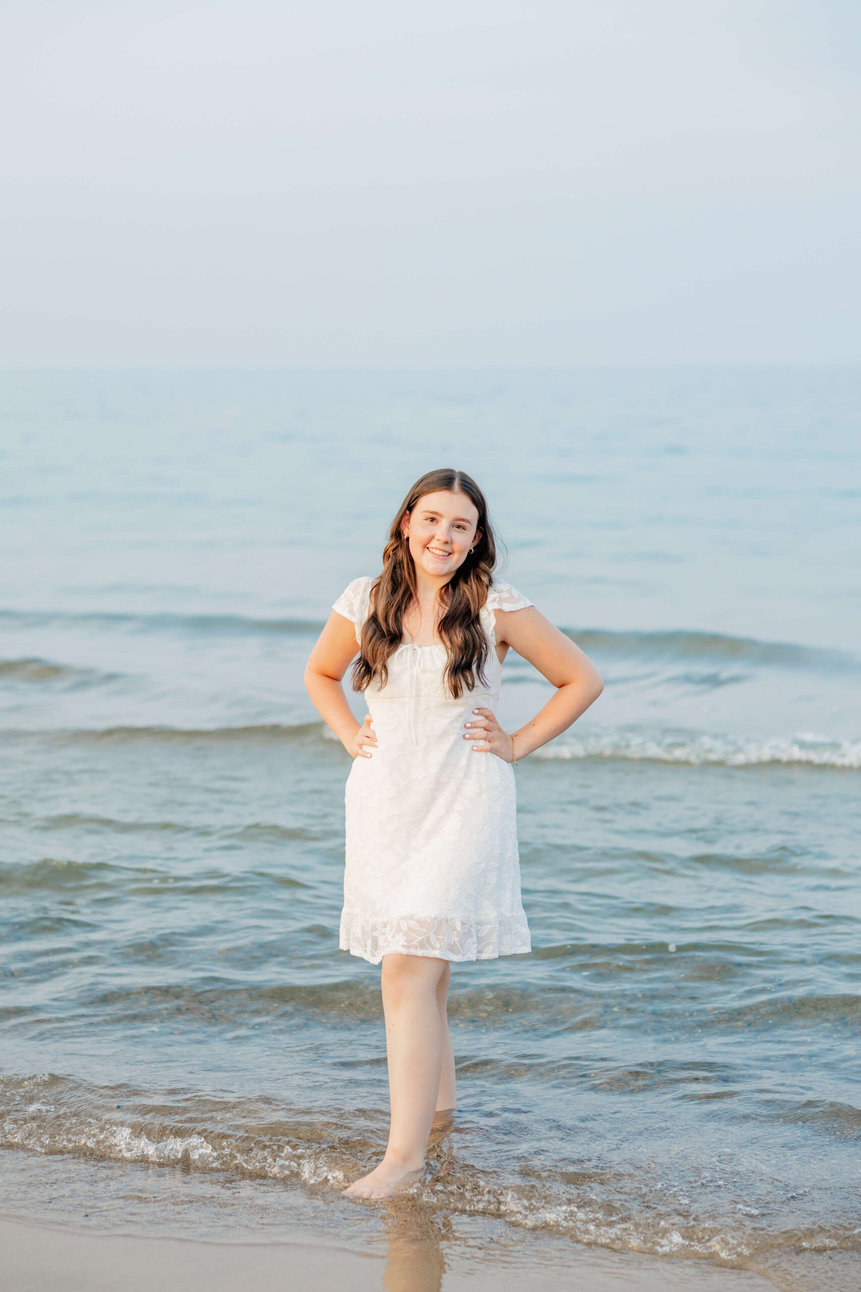 A happy high school senior stands in the water with hands on her hips in a white dress smiling before heading to prom in port huron