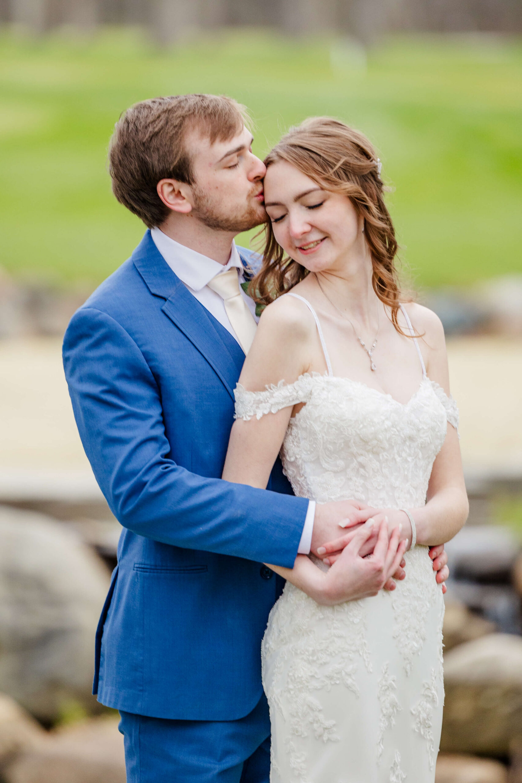A groom kisses his happy bride's forehead by a river at their wyndgate country club wedding