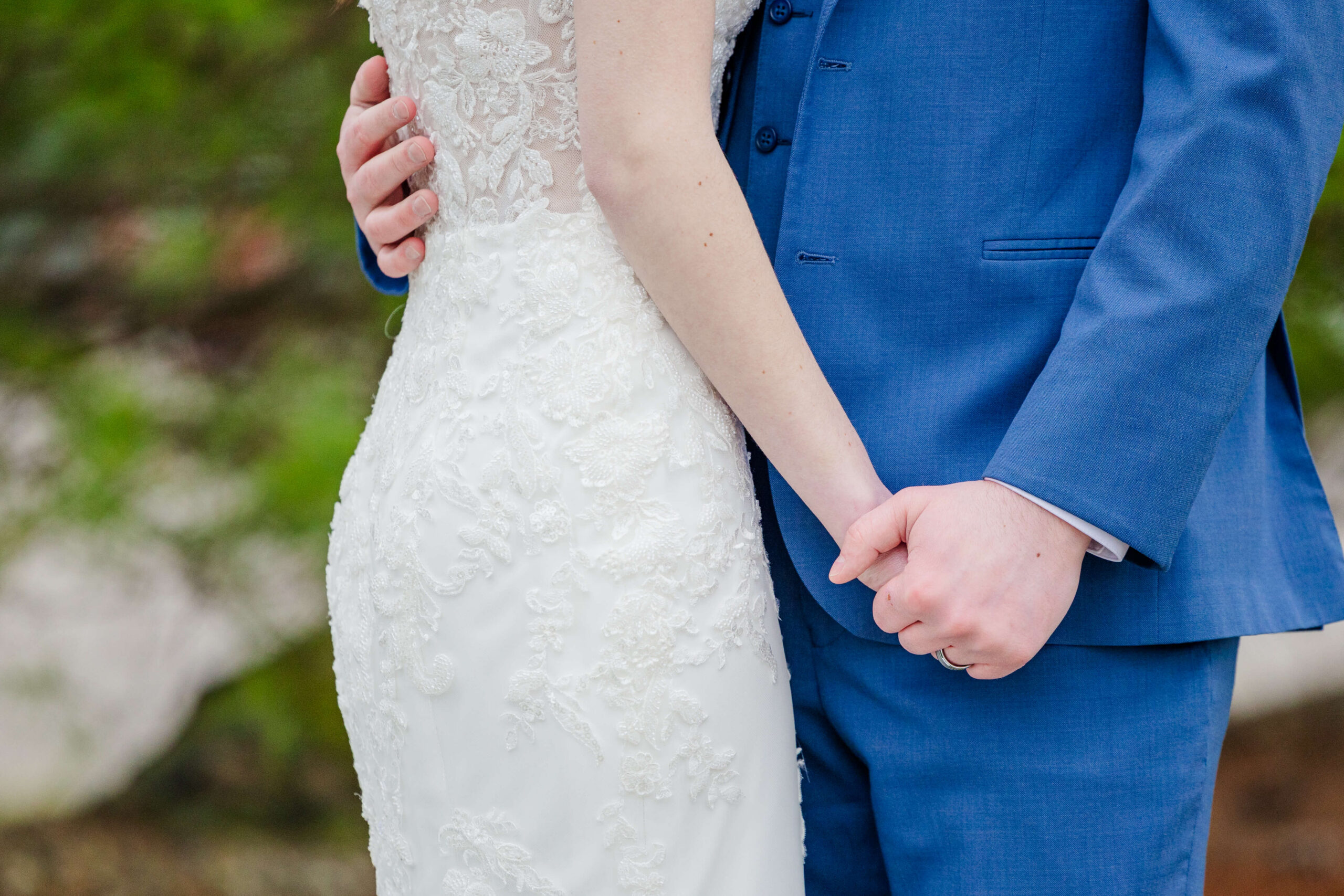 Details of newlyweds holding hands in front of blue suit while standing in garden