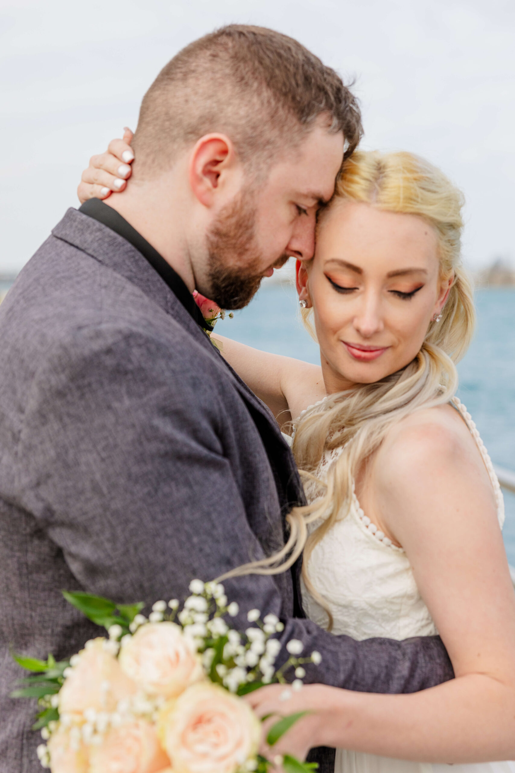 A bride smiles down her shoulder while snuggling her groom on a dock during their Lighthouse Cove wedding