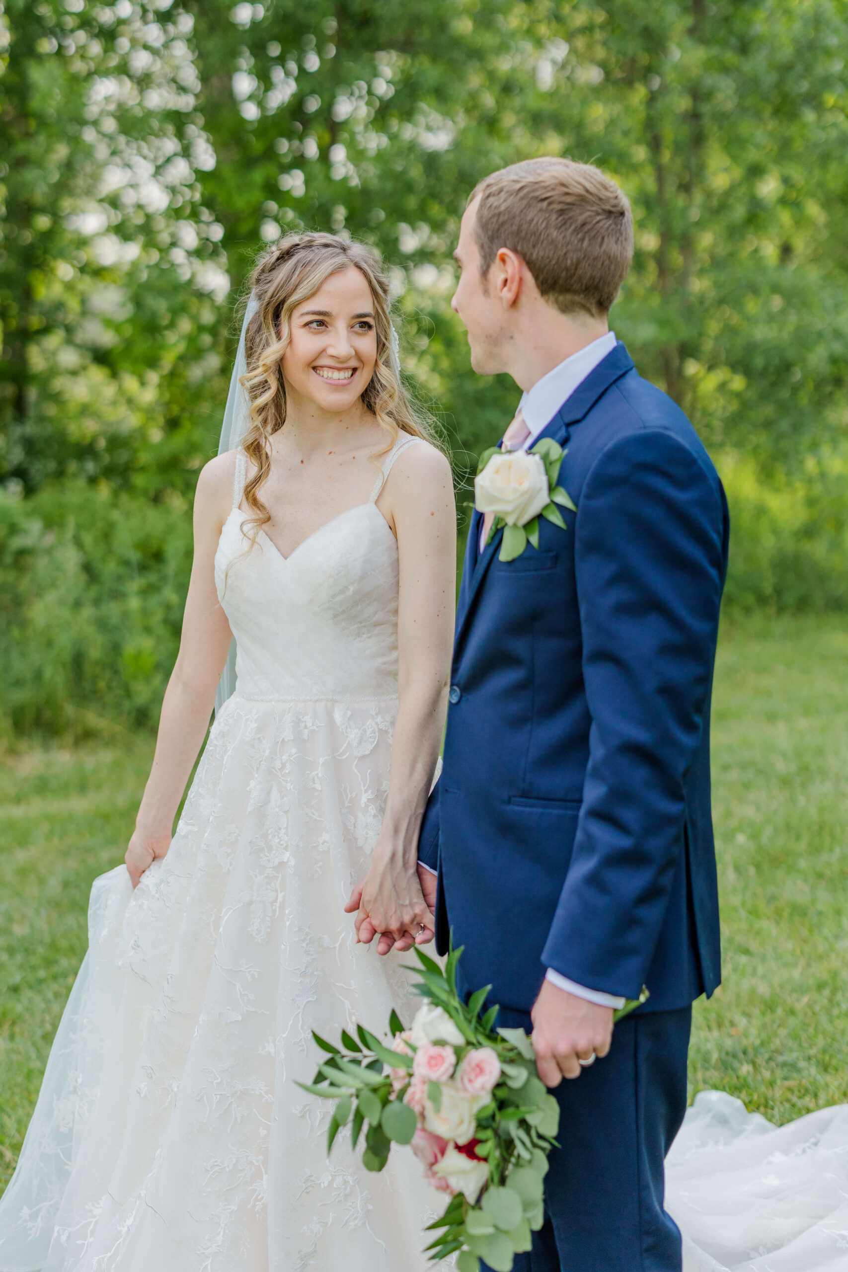 A happy bride smiles at her groom while walking holding hands in a lawn in her lace gown and blue suit