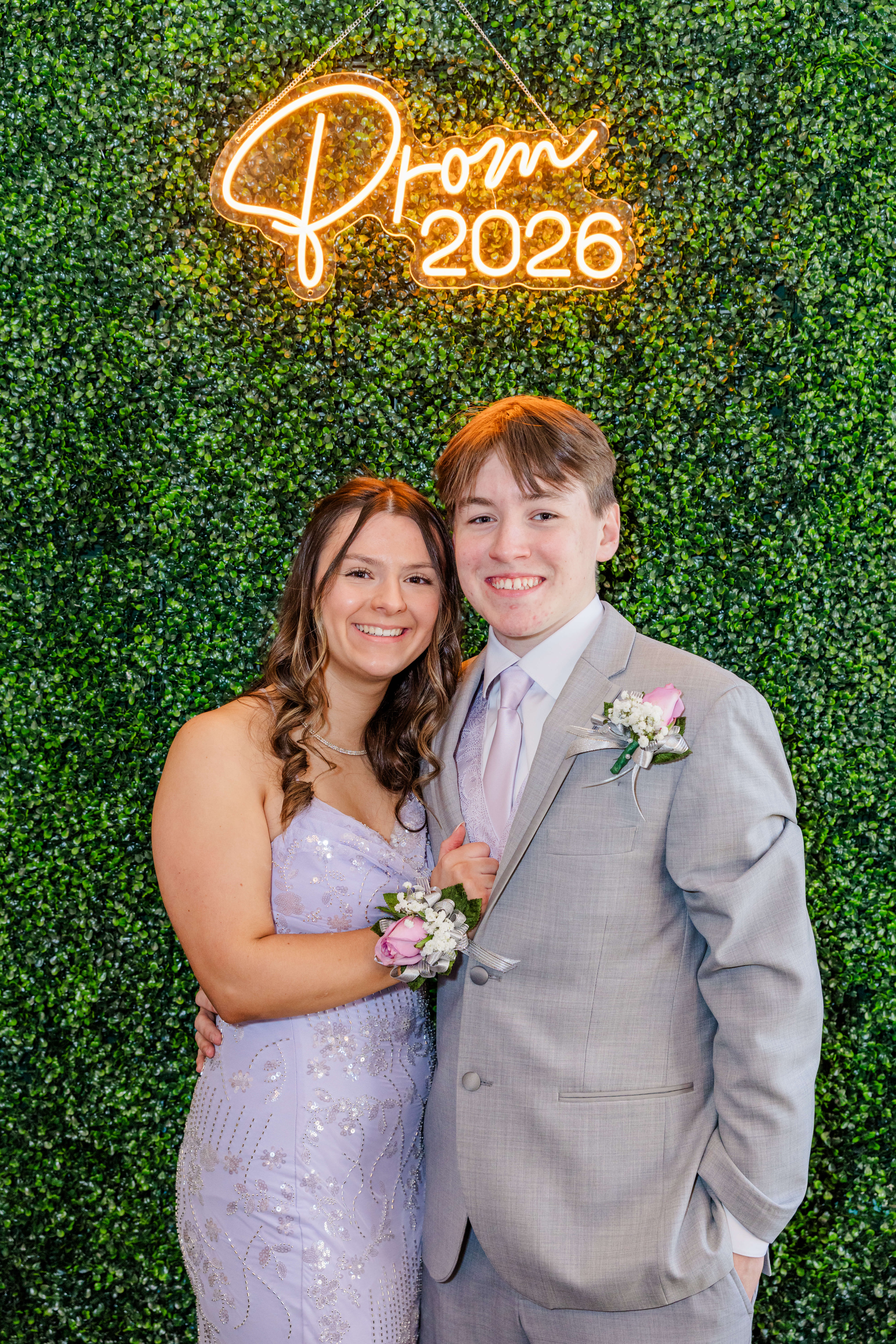A happy prom couple in purple stand smiling under a neon prom sign