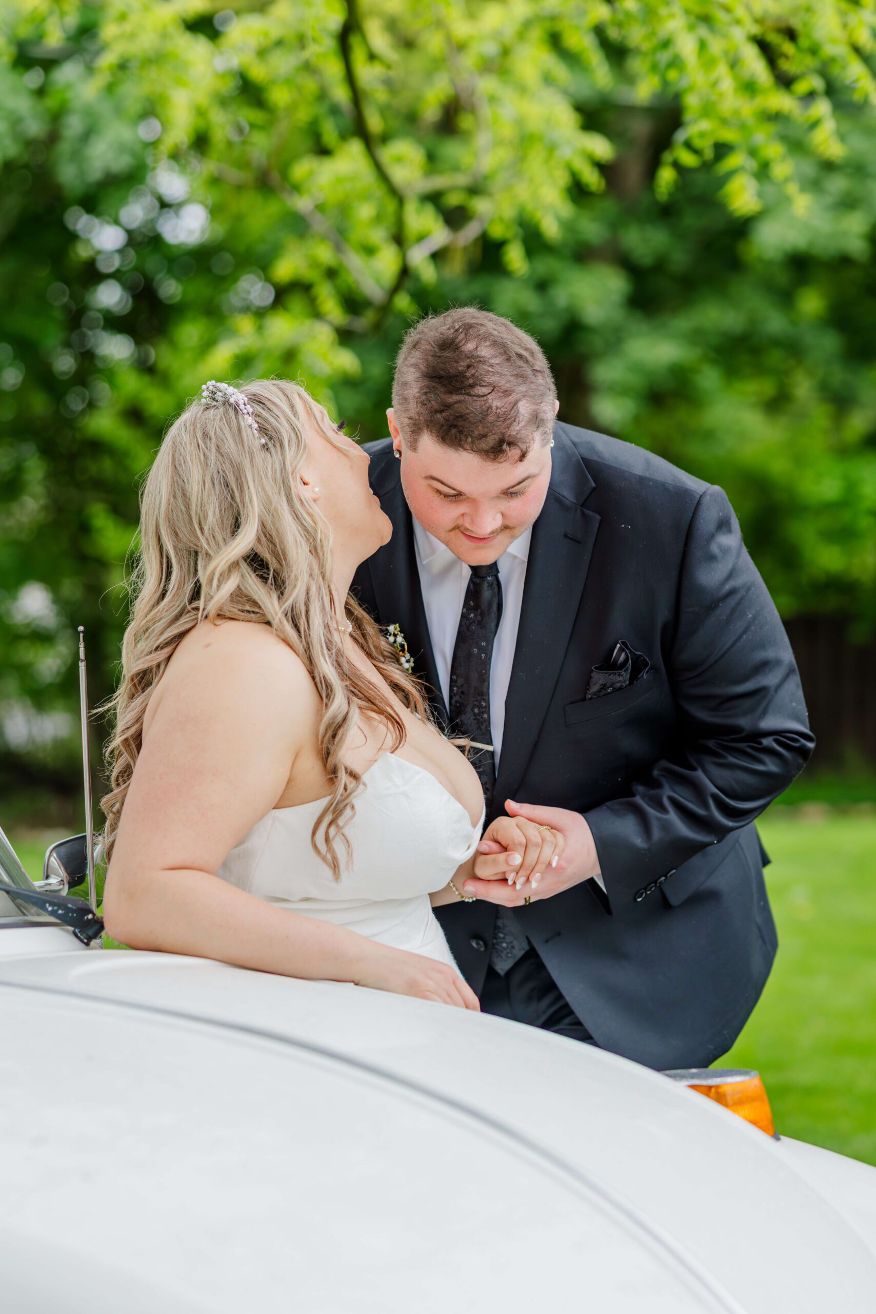 A bride whispers into her groom's ear as they hold hans leaning against a vintage white car