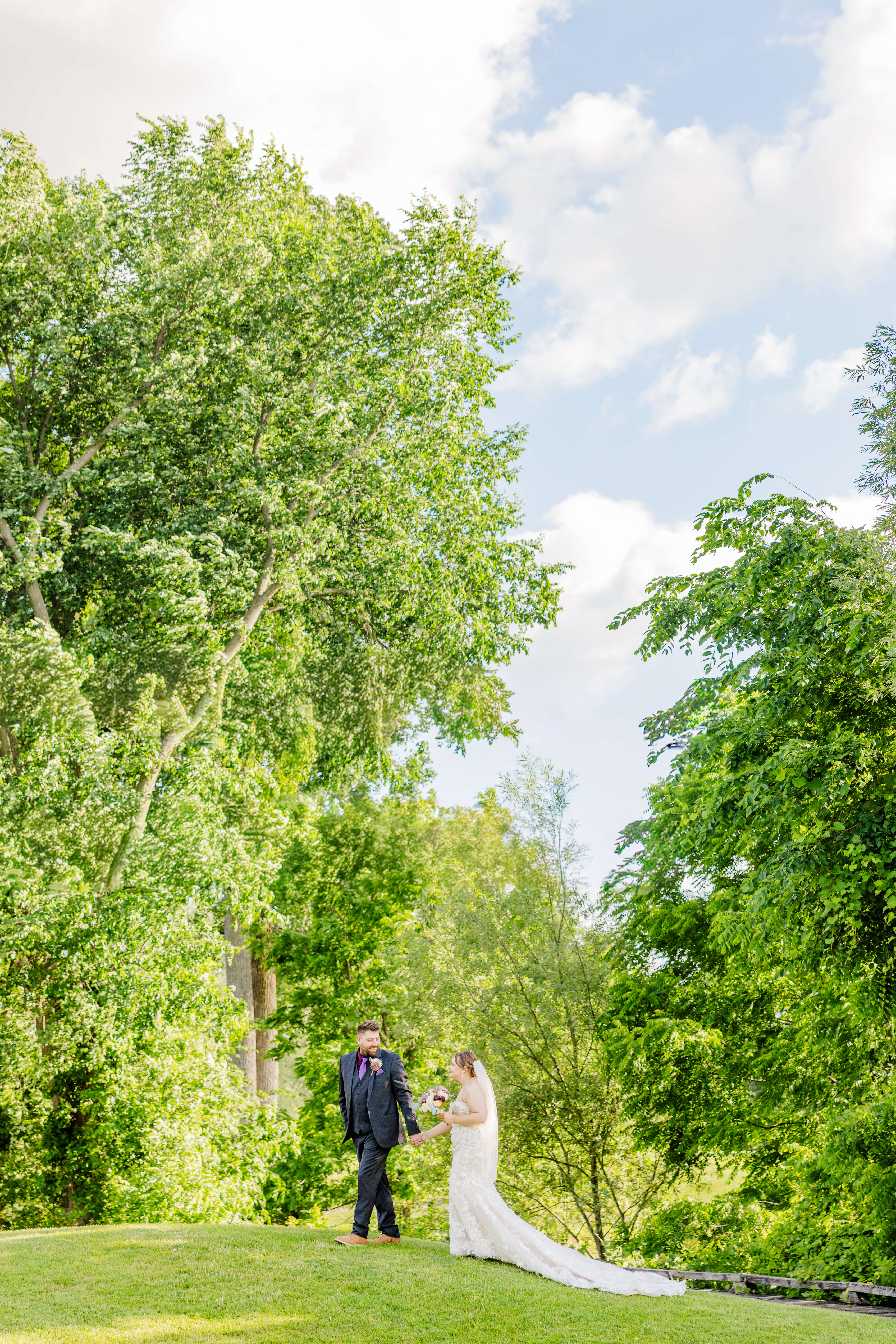 A groom in grey and purple walks his bride by the hand through a lawn under trees