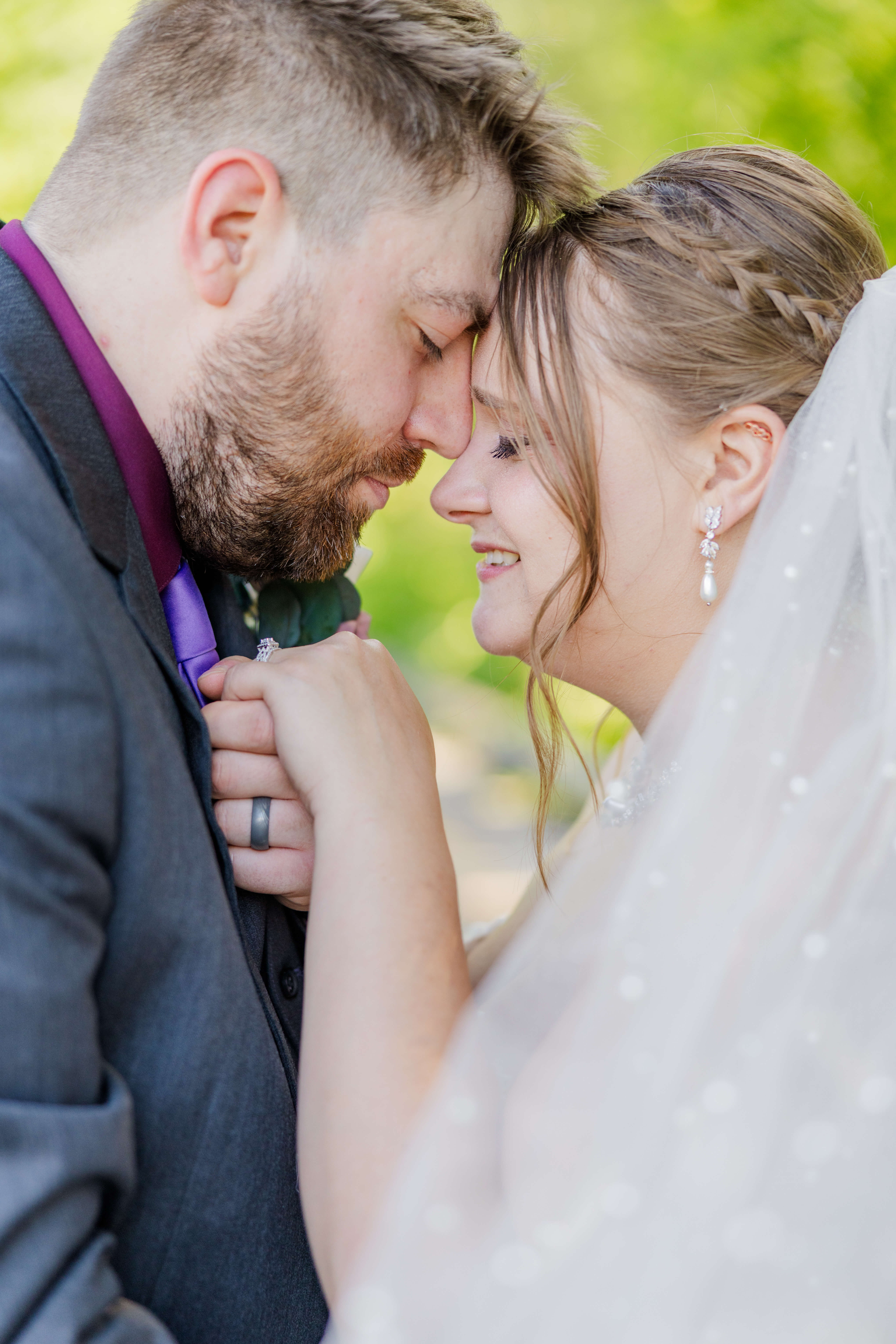 A bride smiles while touching foreheads and holding hands with her groom in grey and purple at their lapeer country club wedding
