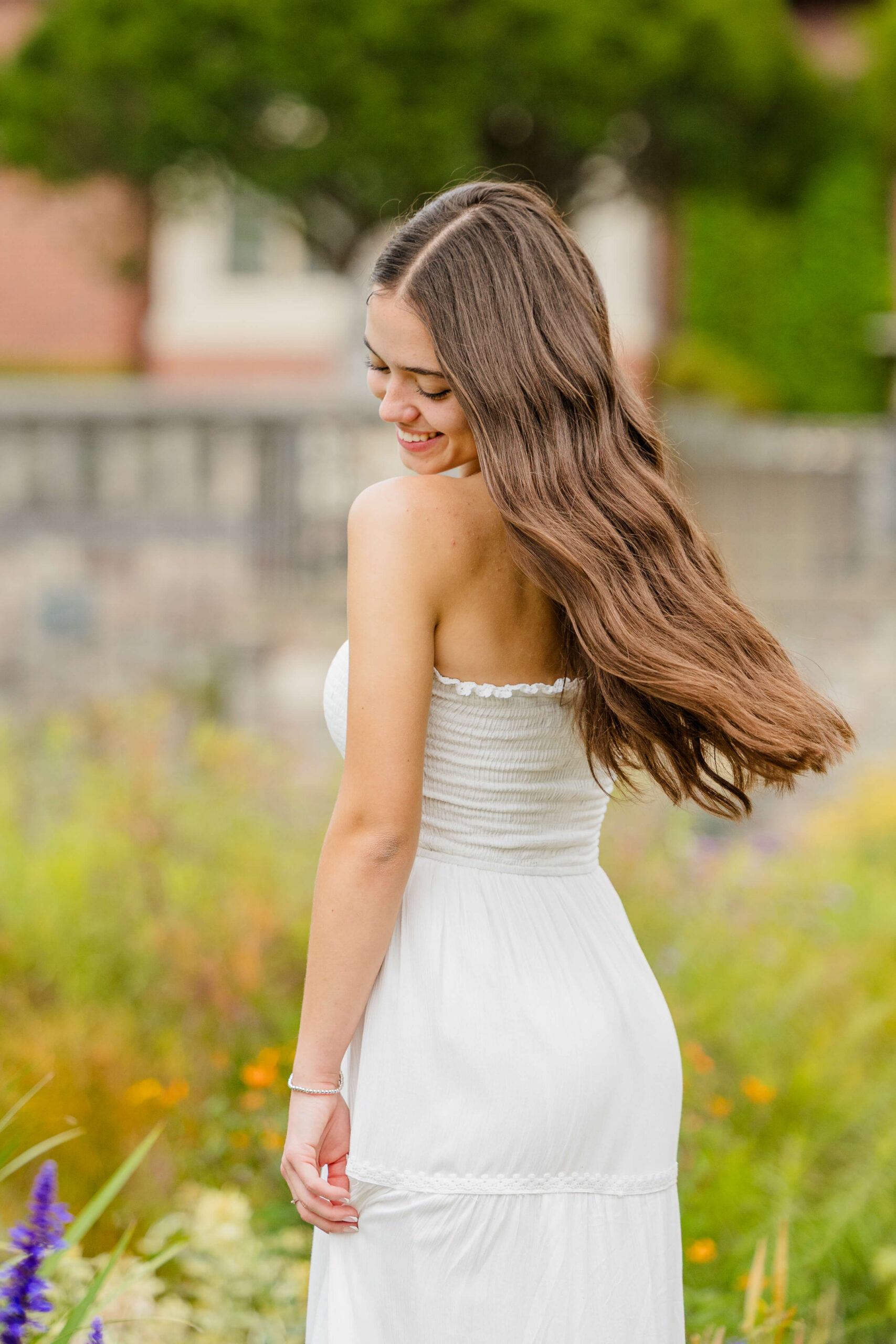 A happy high school senior in a white dress laughs while looking down her shoulder and flipping her hair in a garden