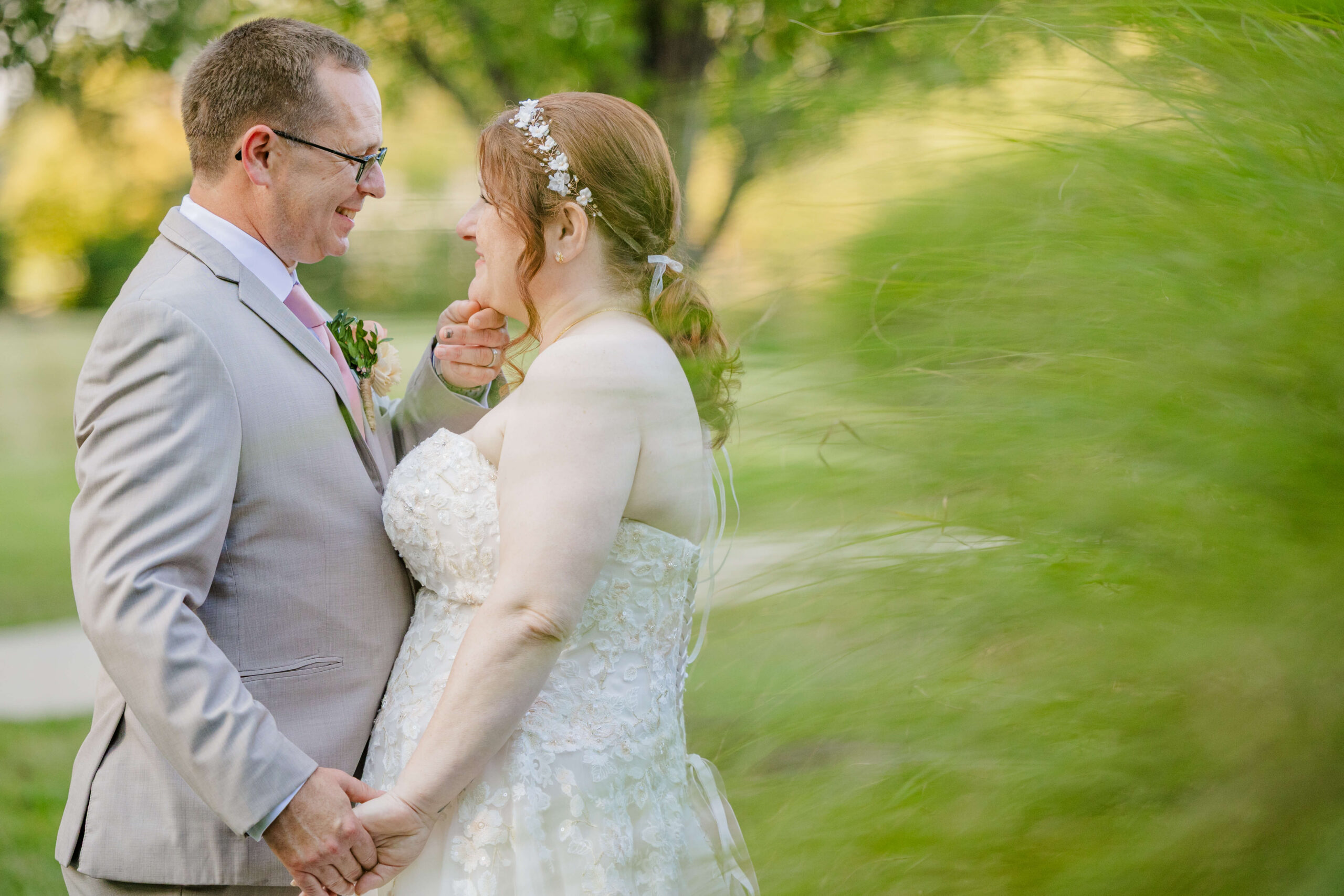 A bride and groom smile at each other in a tan suit and lace gown at sunset holding hands in the gardens of spring meadows country club wedding